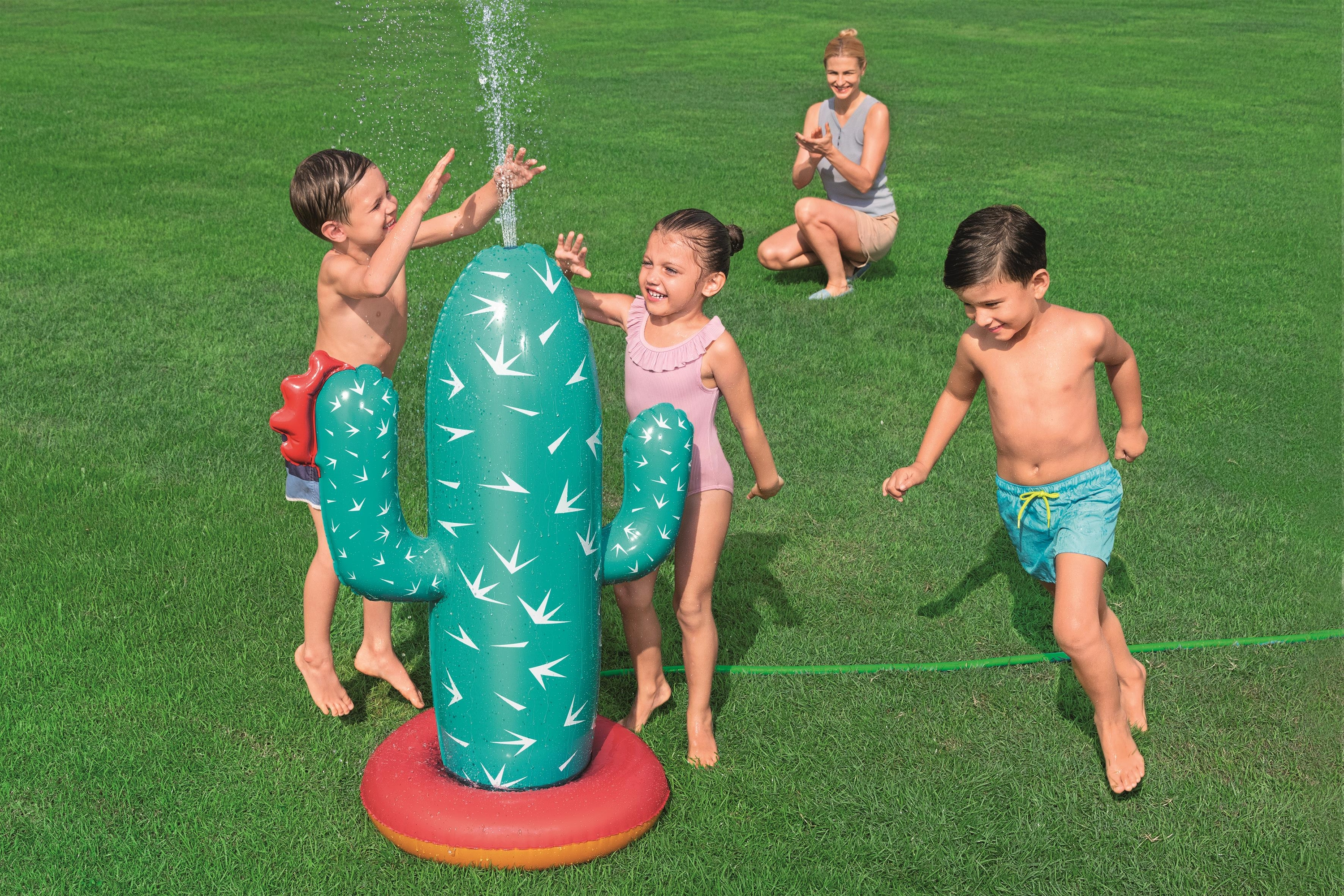 Dzieci bawią się pod zraszaczem kaktusa. Kobieta klaska w tle na zielonym trawniku.