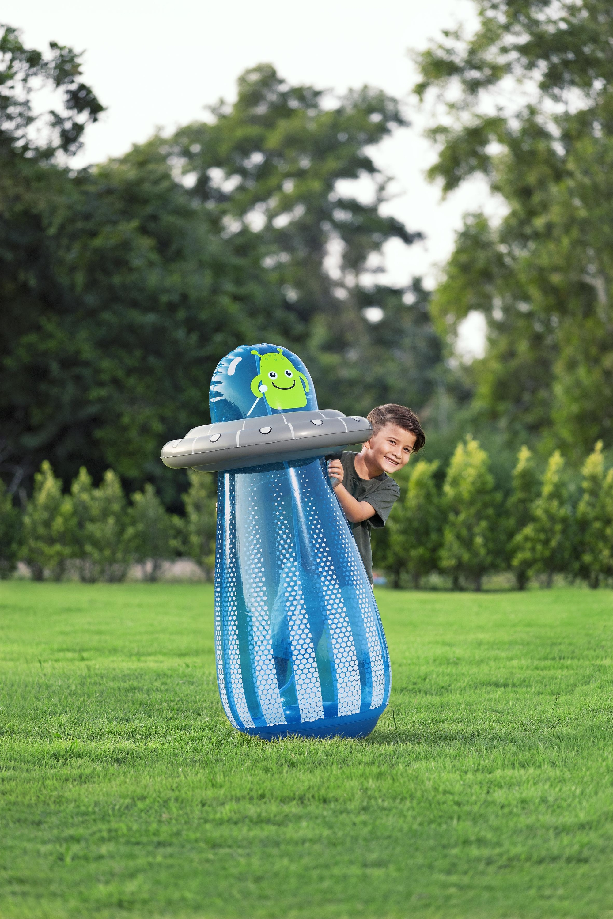 Chłopiec stoi za nadmuchiwaną zabawką statek kosmiczny obcych na trawiastym polu. Zabawka ma zielonego obcego na górze.