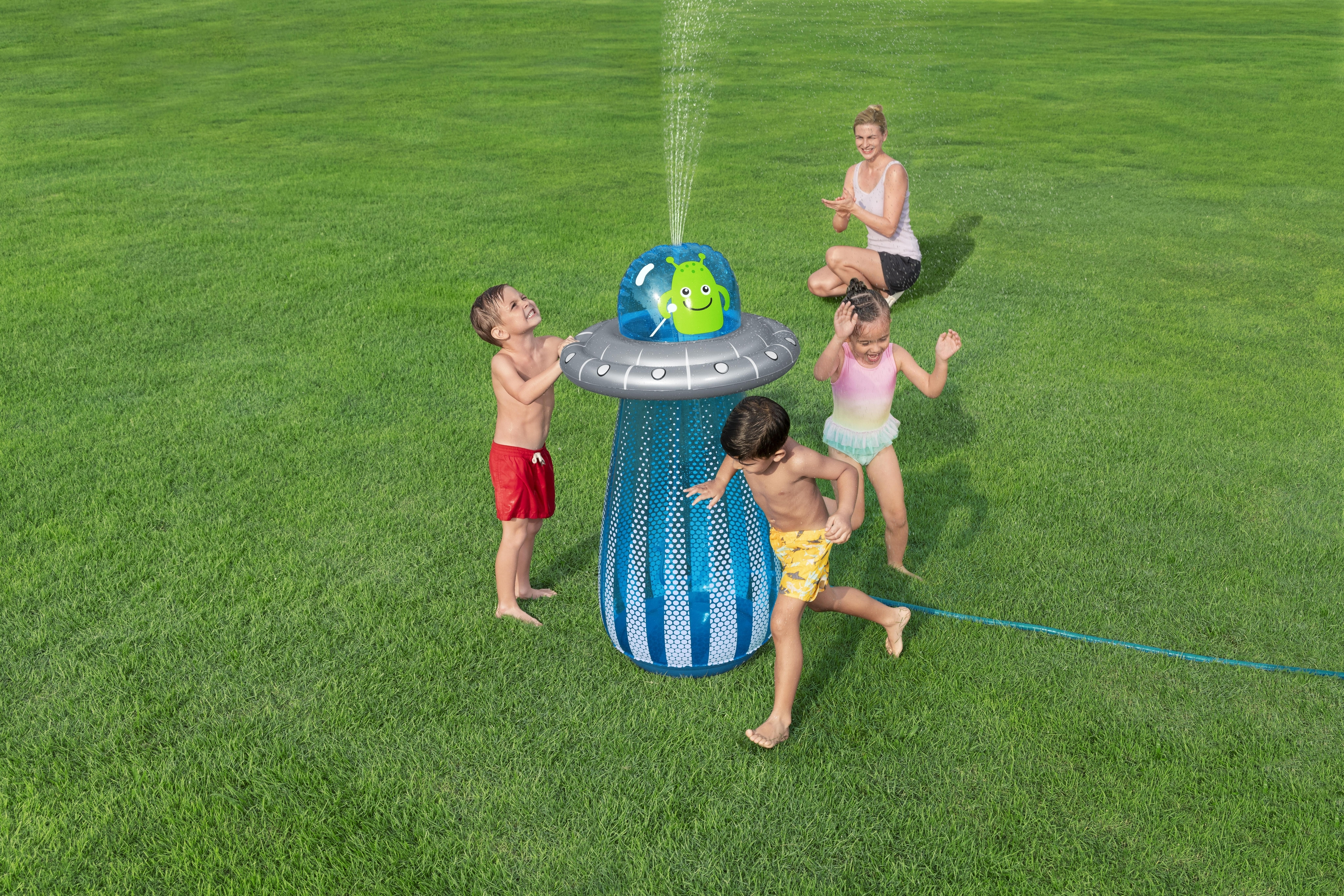 Dzieci bawią się na podwórku z nadmuchiwanym zraszaczem w kształcie statku kosmicznego obcych. Kobieta obserwuje ich z trawy.