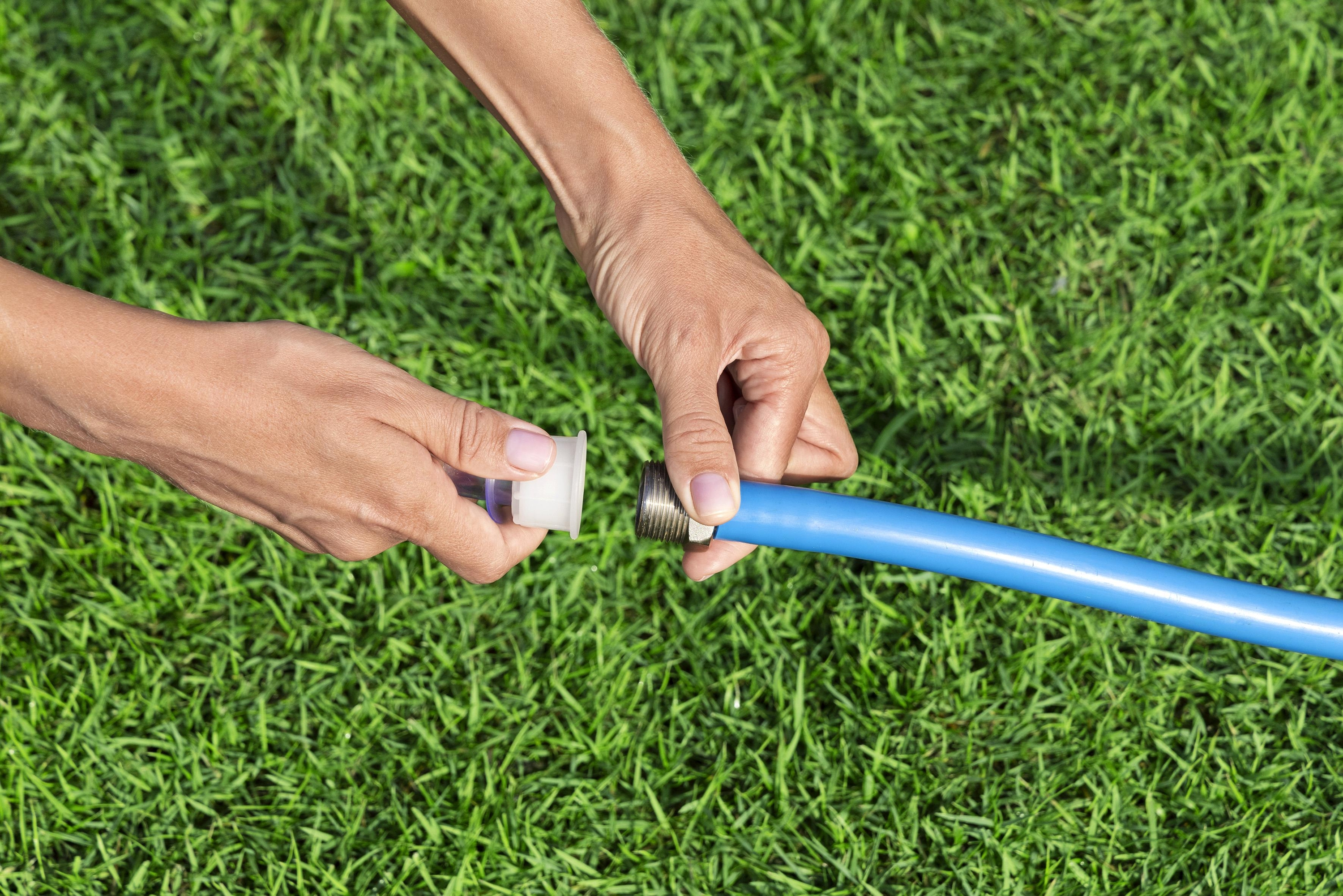 Ręce łączą niebieski wąż z białym łącznikiem na zielonej trawie.