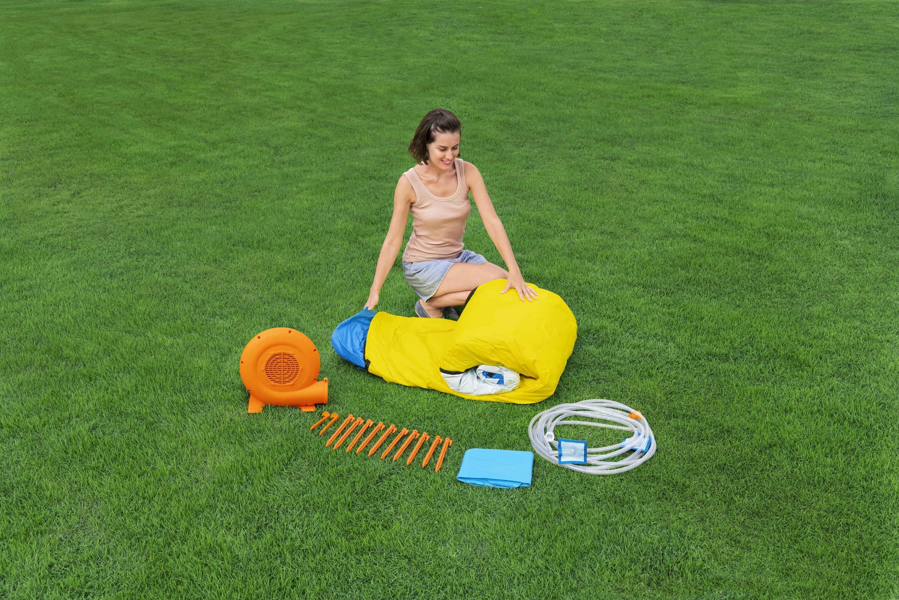 Kobieta rozpakowuje zjeżdżalnię wodną. Pomarańczowa dmuchawa, kołki i niebieska mata w pobliżu.