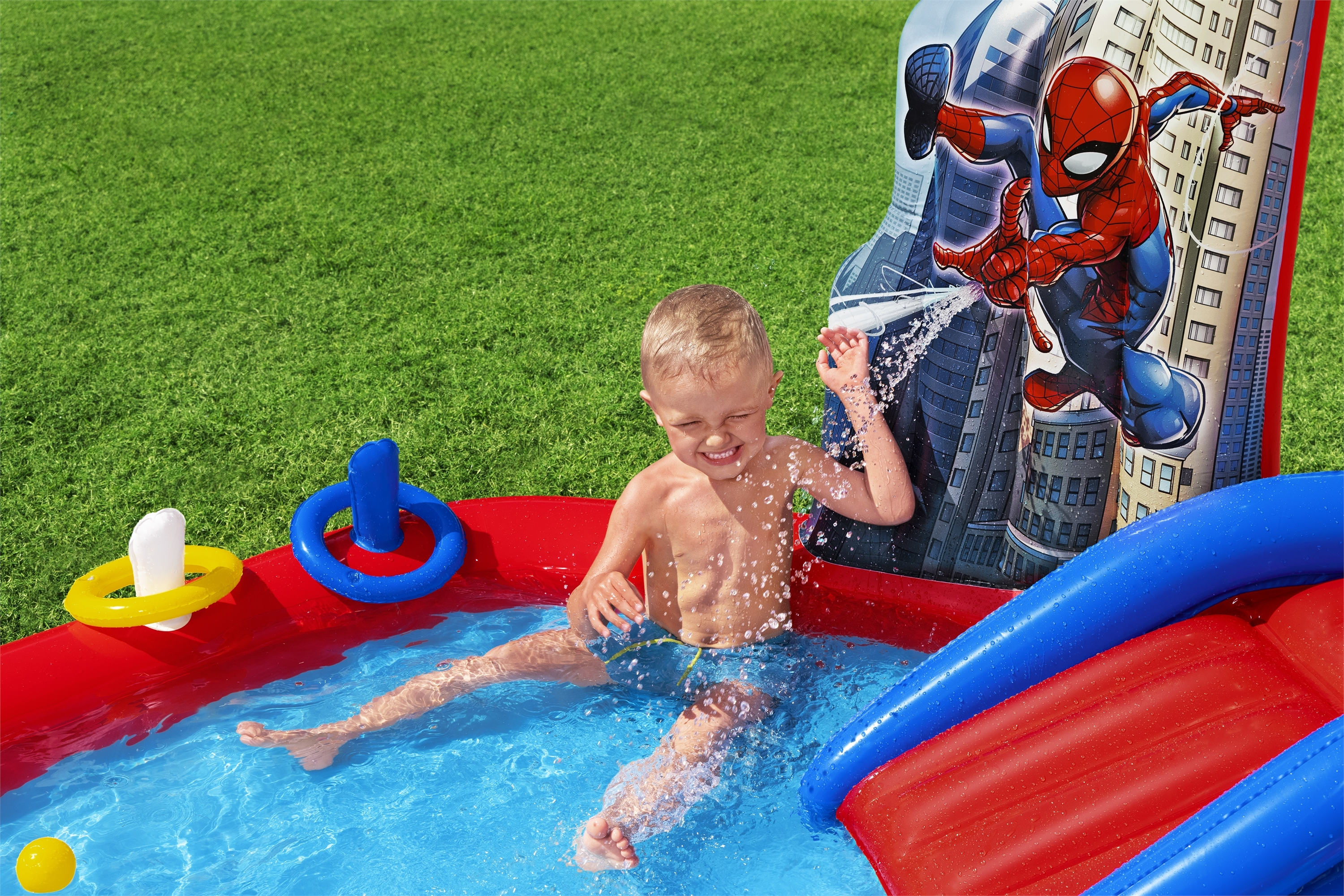 Mały chłopiec bawi się w basenie o tematyce Spider-Mana ze zjeżdżalnią. Basen ma czerwoną i niebieską kolorystykę.