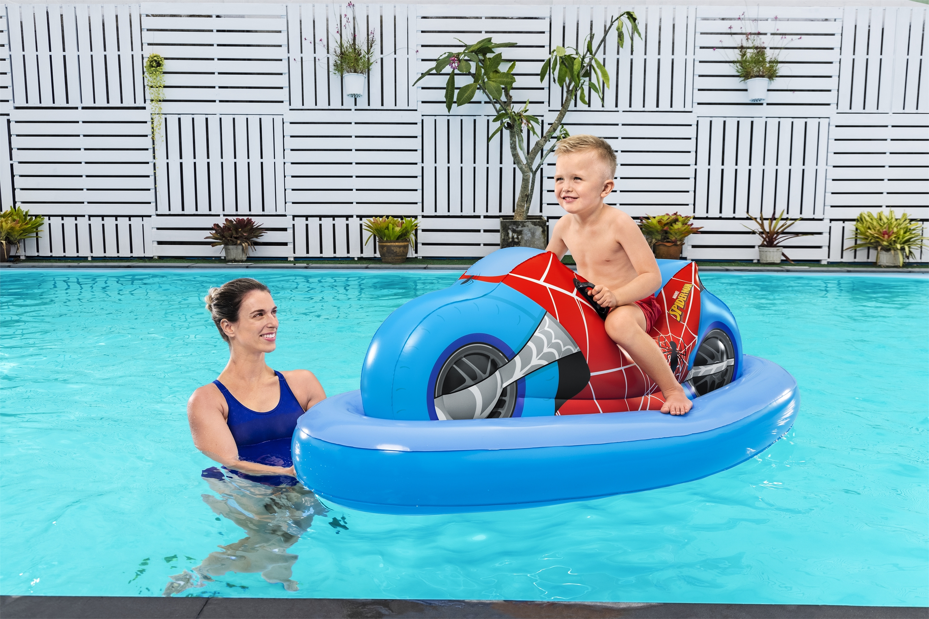Ein Junge auf einem blauen Spielzeugmotorrad in einem Pool, eine Frau schaut zu. Weißer Zaun.
