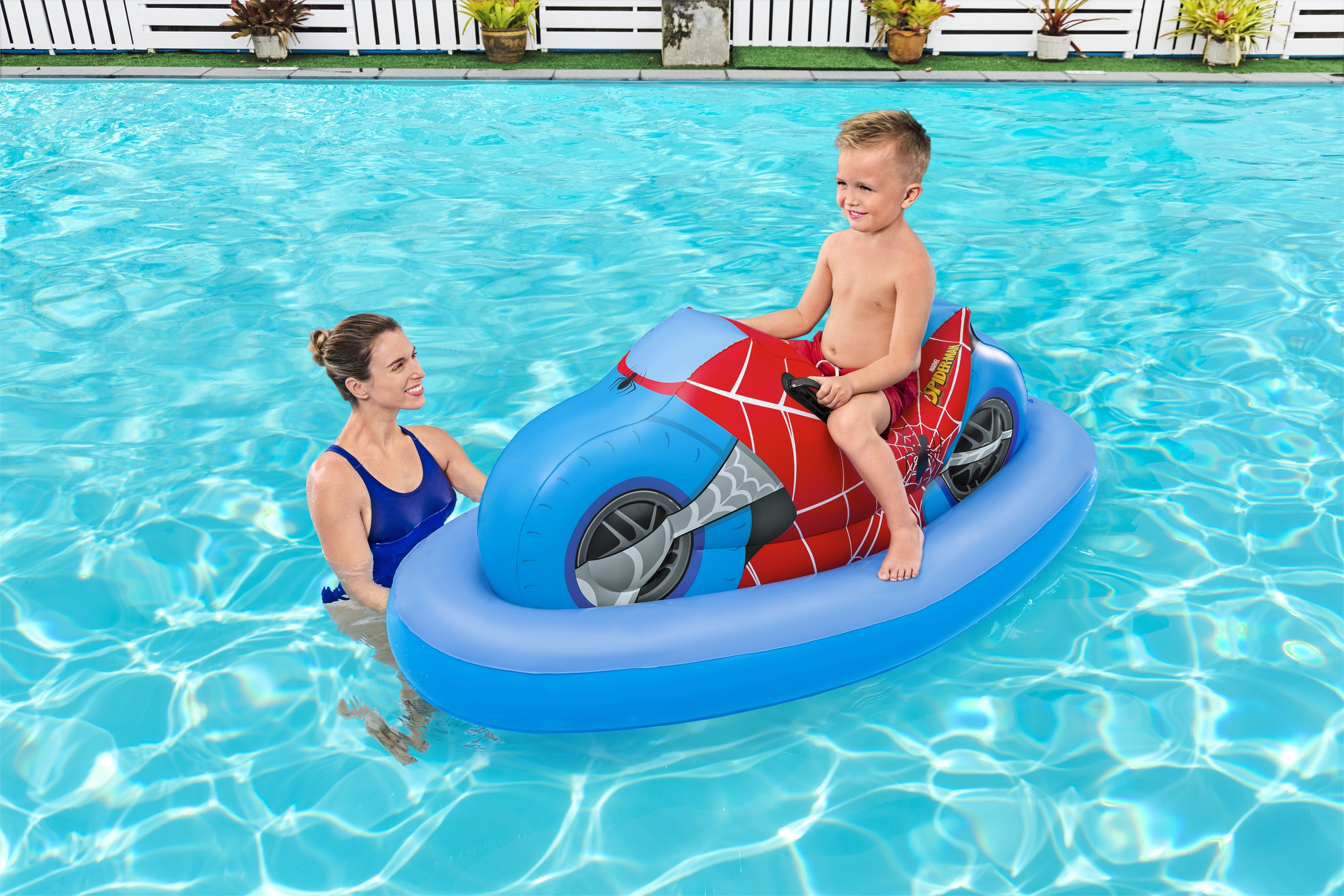 Ein Junge sitzt auf einem aufblasbaren Jet-Ski im Spider-Man-Design in einem Pool, eine Frau in der Nähe.