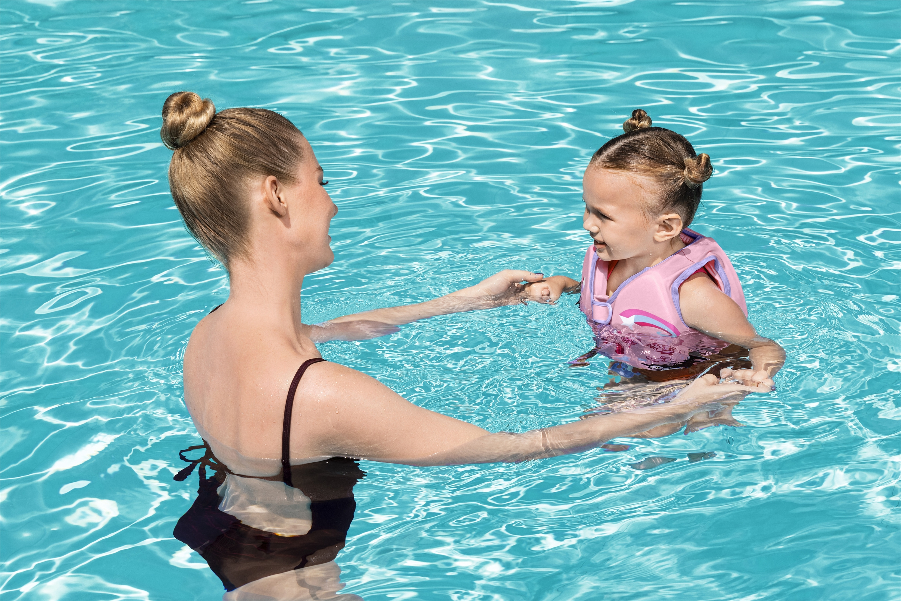 Donna in costume che tiene le mani a una bambina con gilet rosa in piscina.