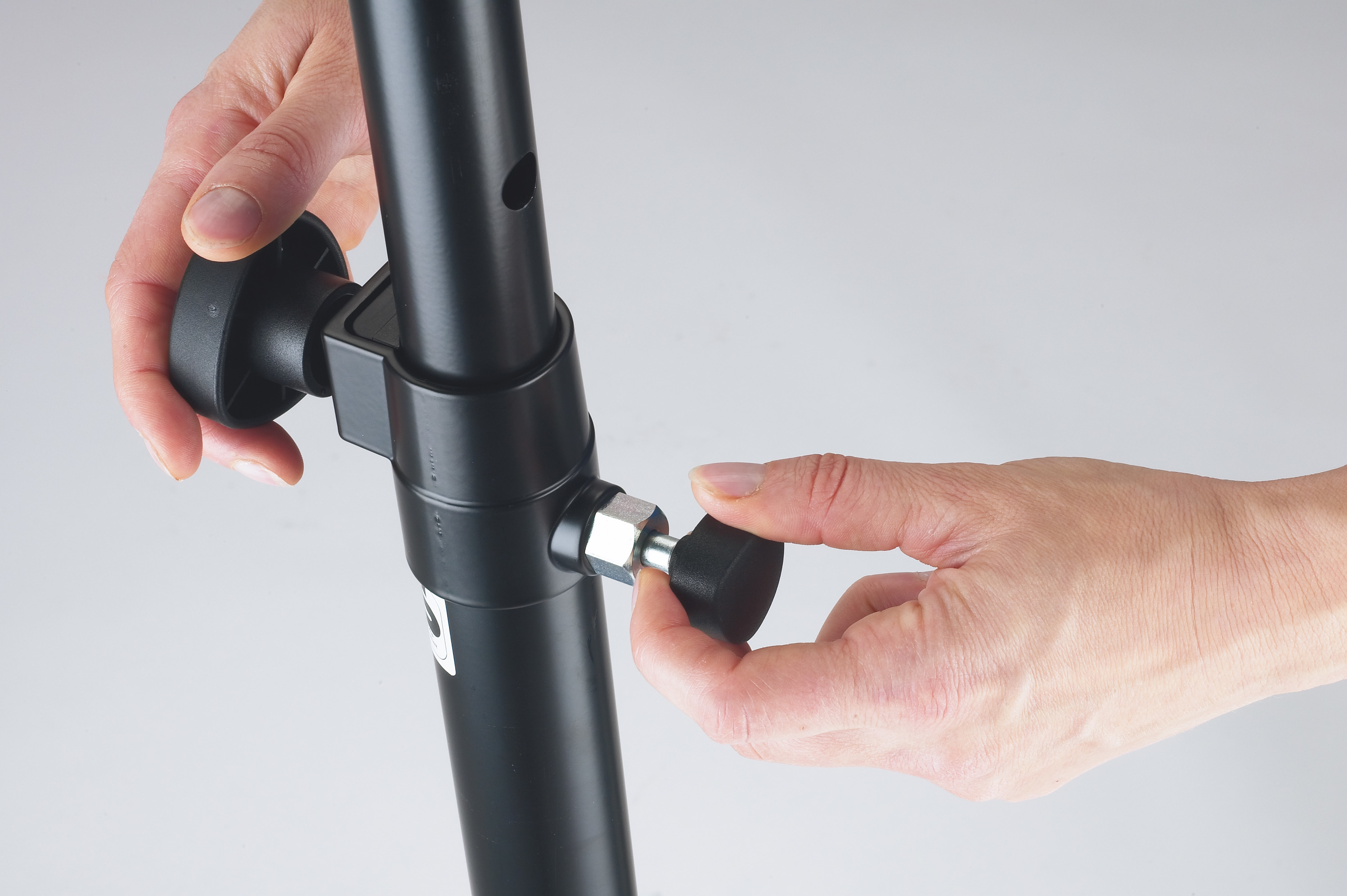 Dłonie regulujące czarny statyw za pomocą pokrętła na szarym tle.