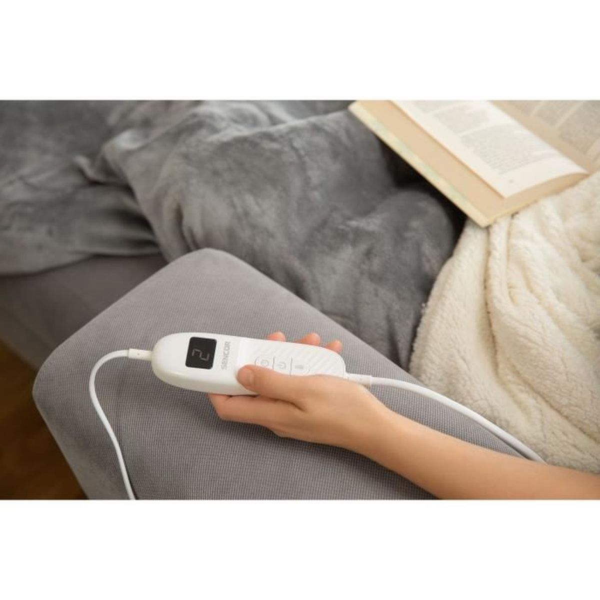 Osoba trzymająca biały kontroler z wyświetlaczem, leżąca na szarym kocu.