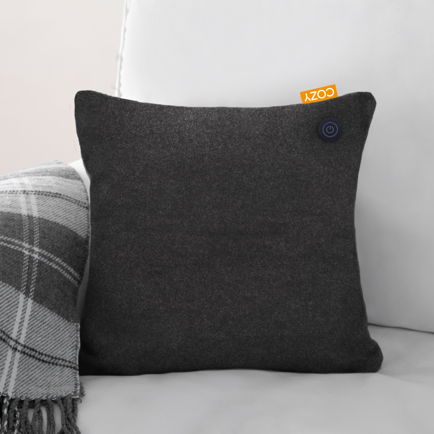 Donkergrijs kussen op witte bank, COZY-label en blauwe knop. Geruite deken zichtbaar.