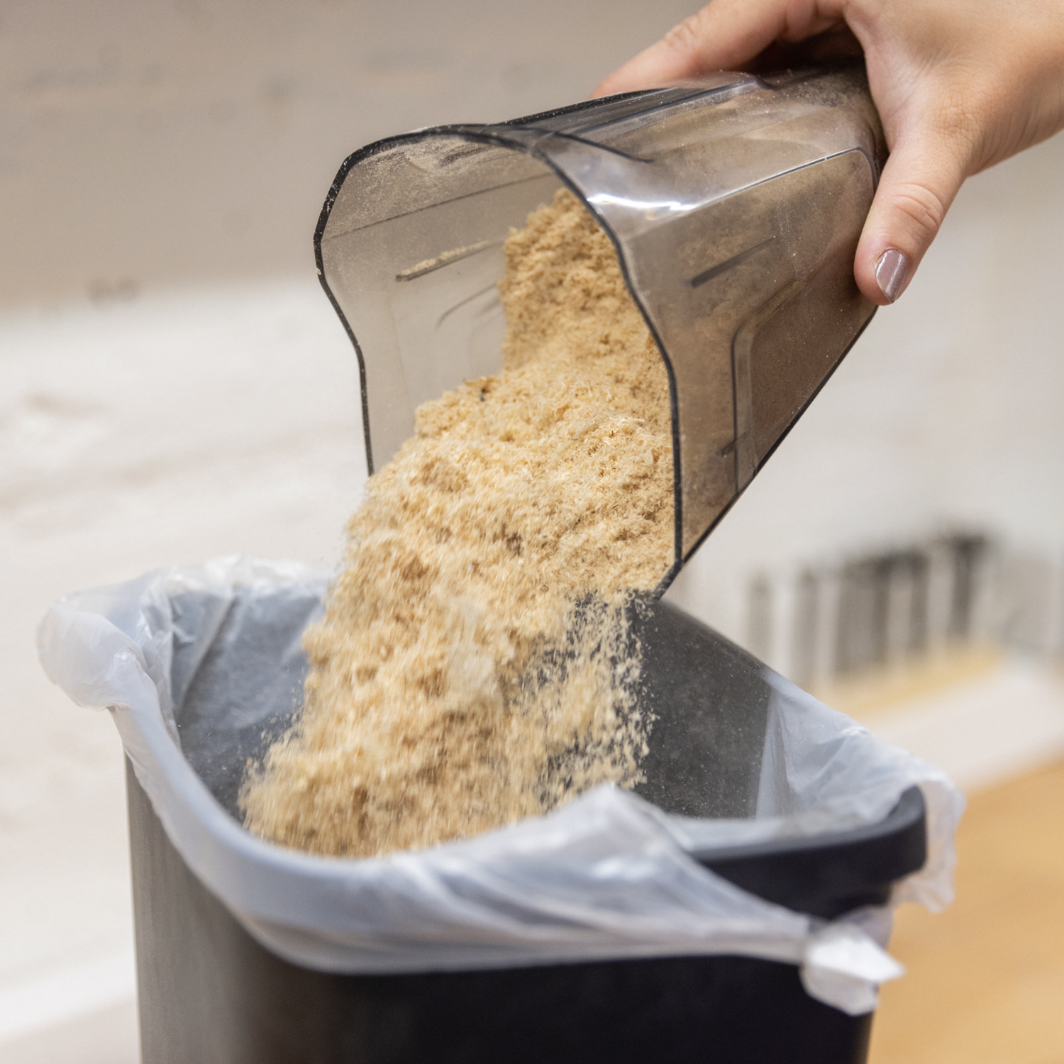 A hand pours sawdust into a black trash can with a bag. The background is a room.