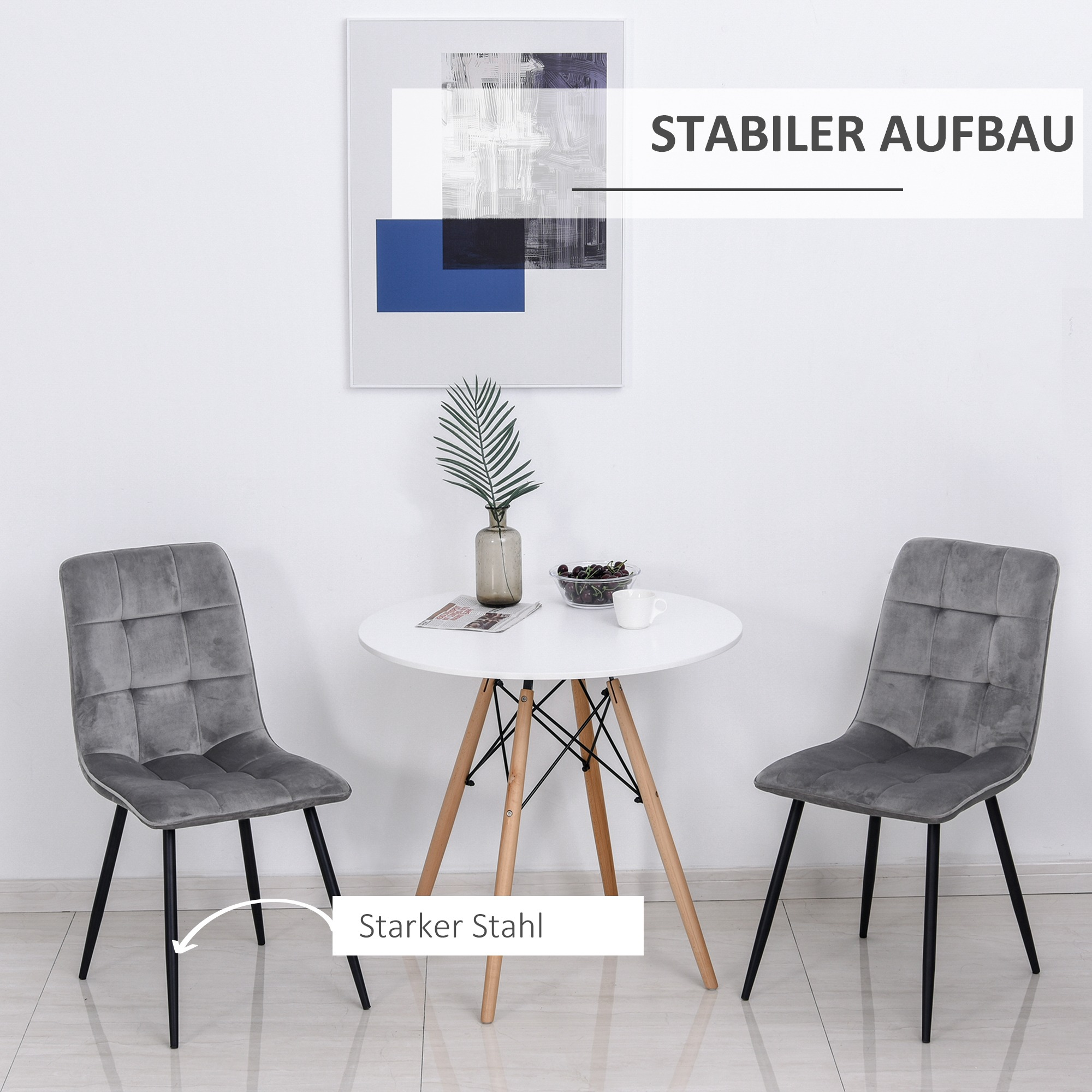 Zwei graue Stühle mit schwarzen Beinen und ein runder weißer Tisch, mit Pflanze und Bild.