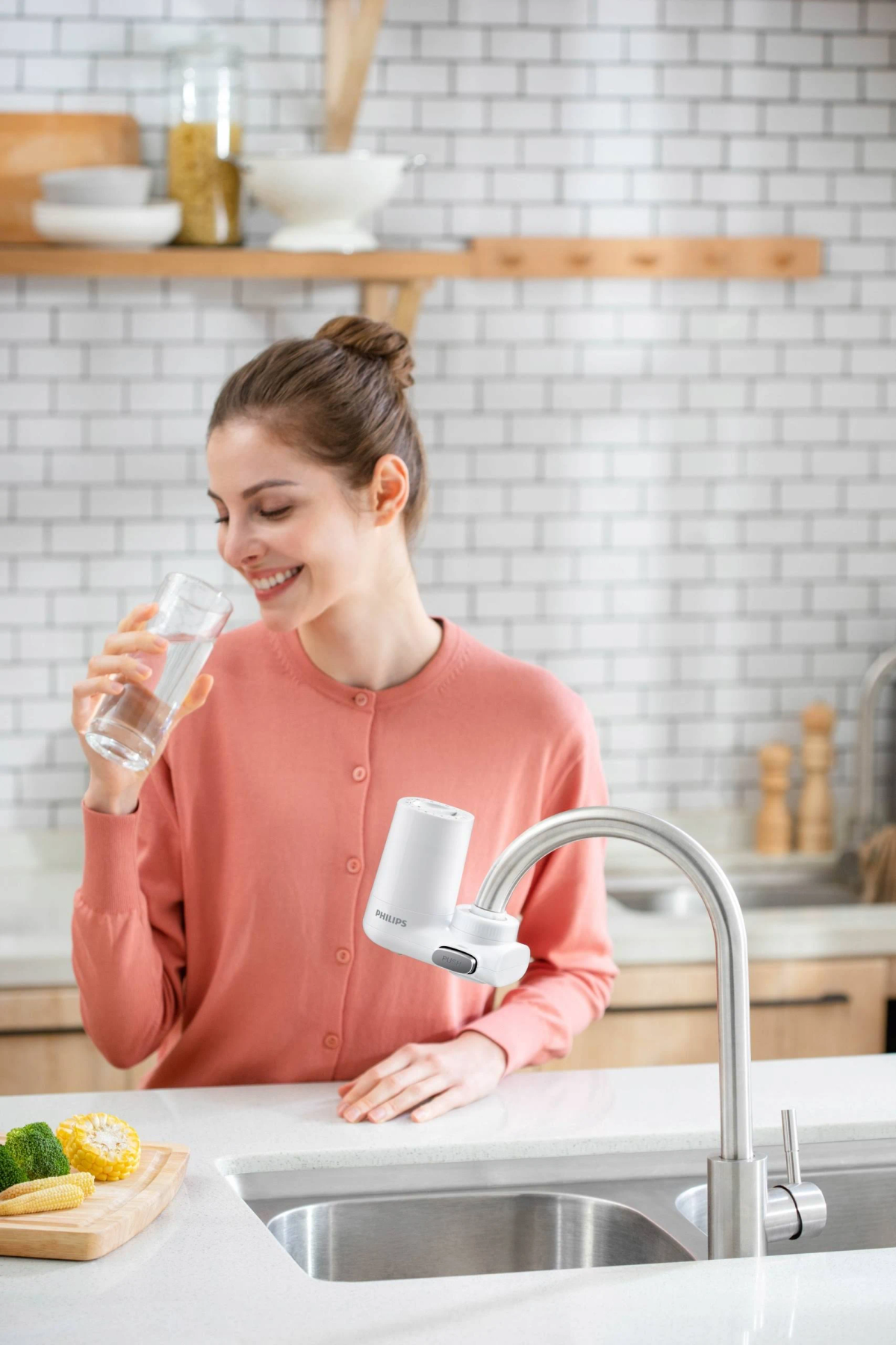 Kobieta pije wodę ze szklanki obok białego filtra wody na kranie w kuchni.