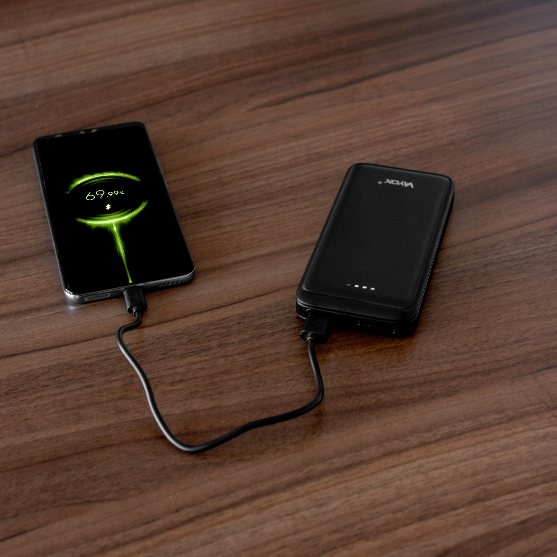 Czarny telefon ładujący czarny power bank. Telefon pokazuje 69,99% naładowania. Drewniana powierzchnia.