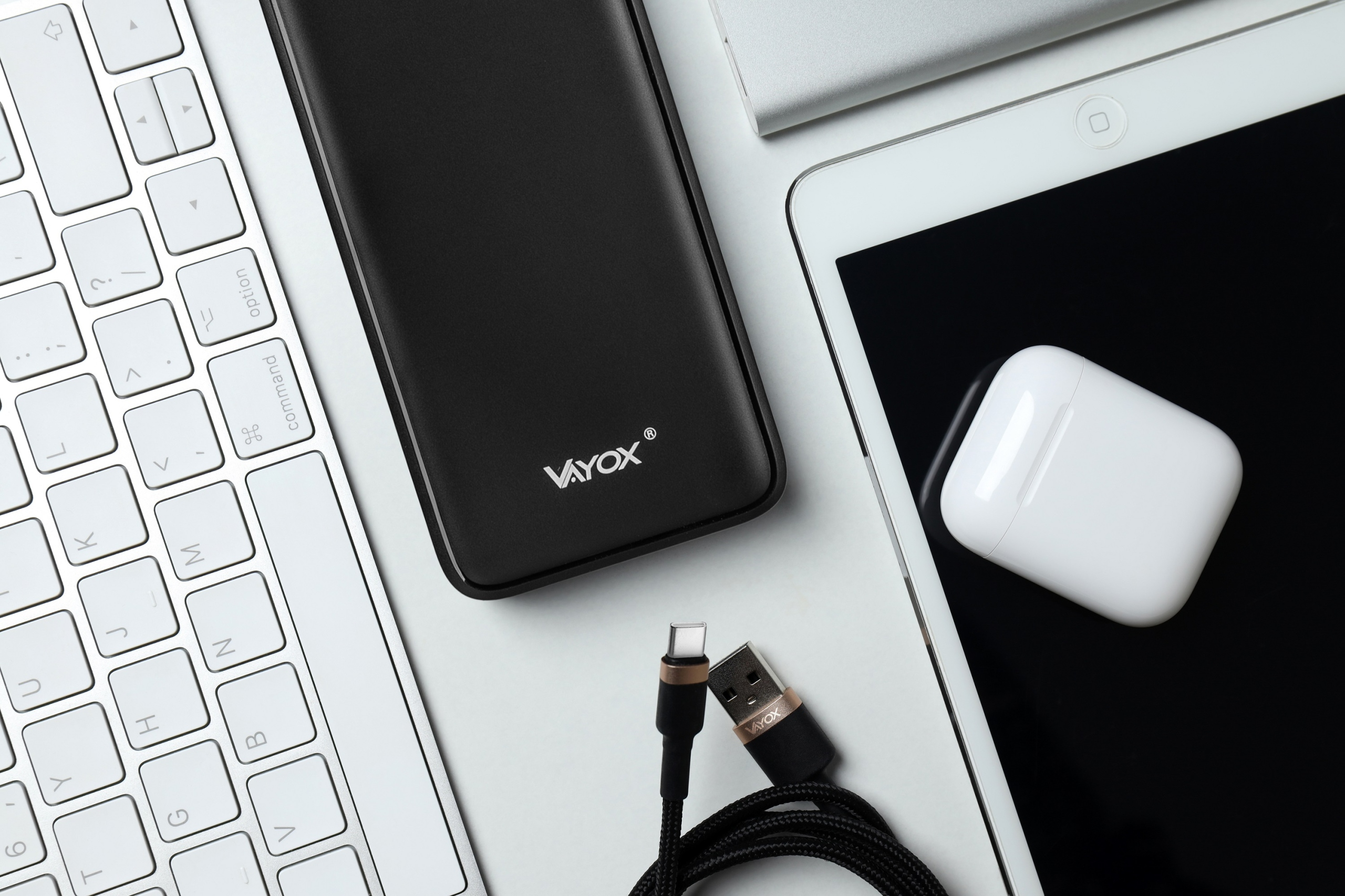 Widok z góry na czarny power bank, biały iPad, AirPods, klawiaturę i kabel USB.