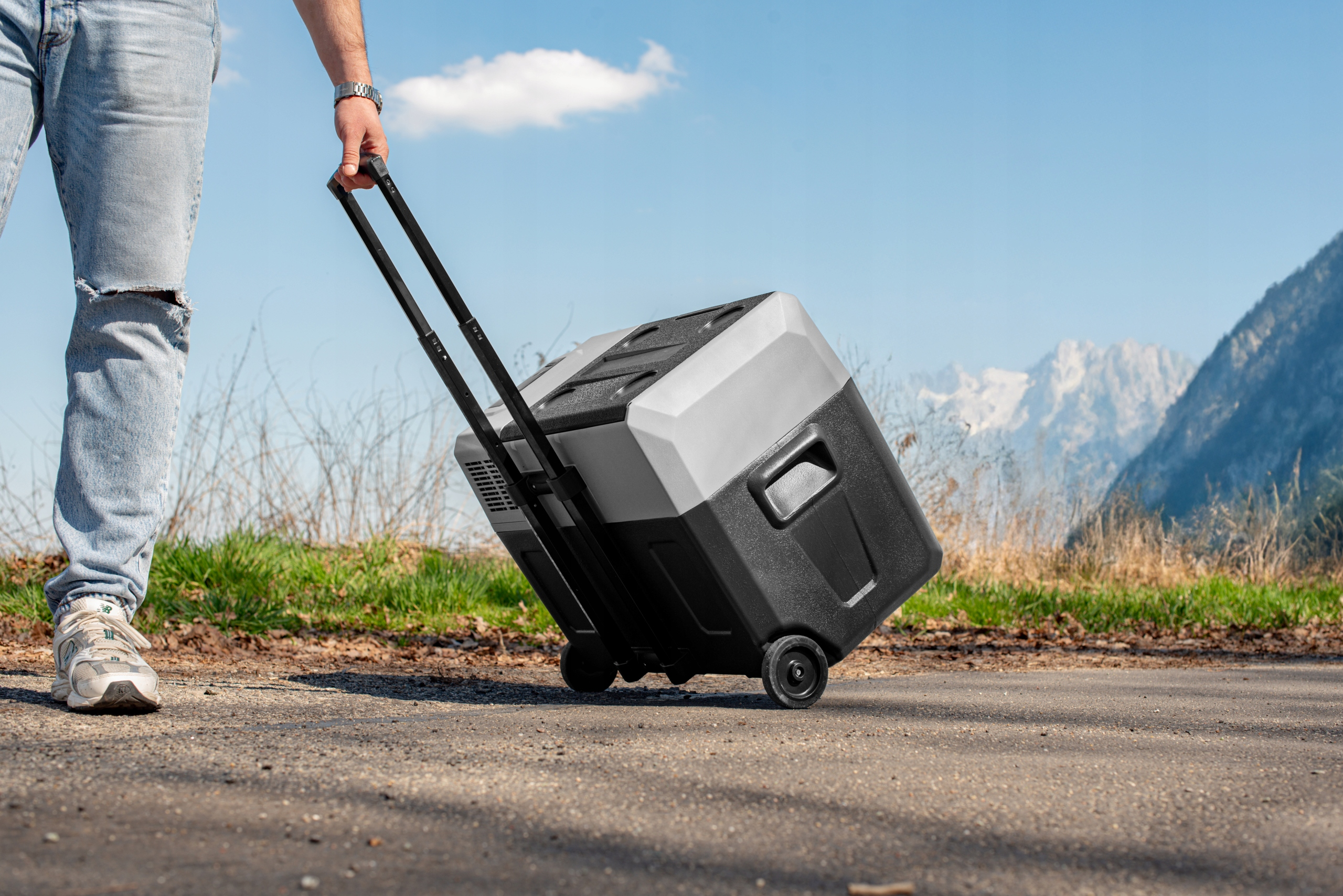 Osoba ciągnąca przenośną chłodziarkę. Mężczyzna w dżinsach i trampkach ciągnie czarno-szarą chłodziarkę na kółkach.