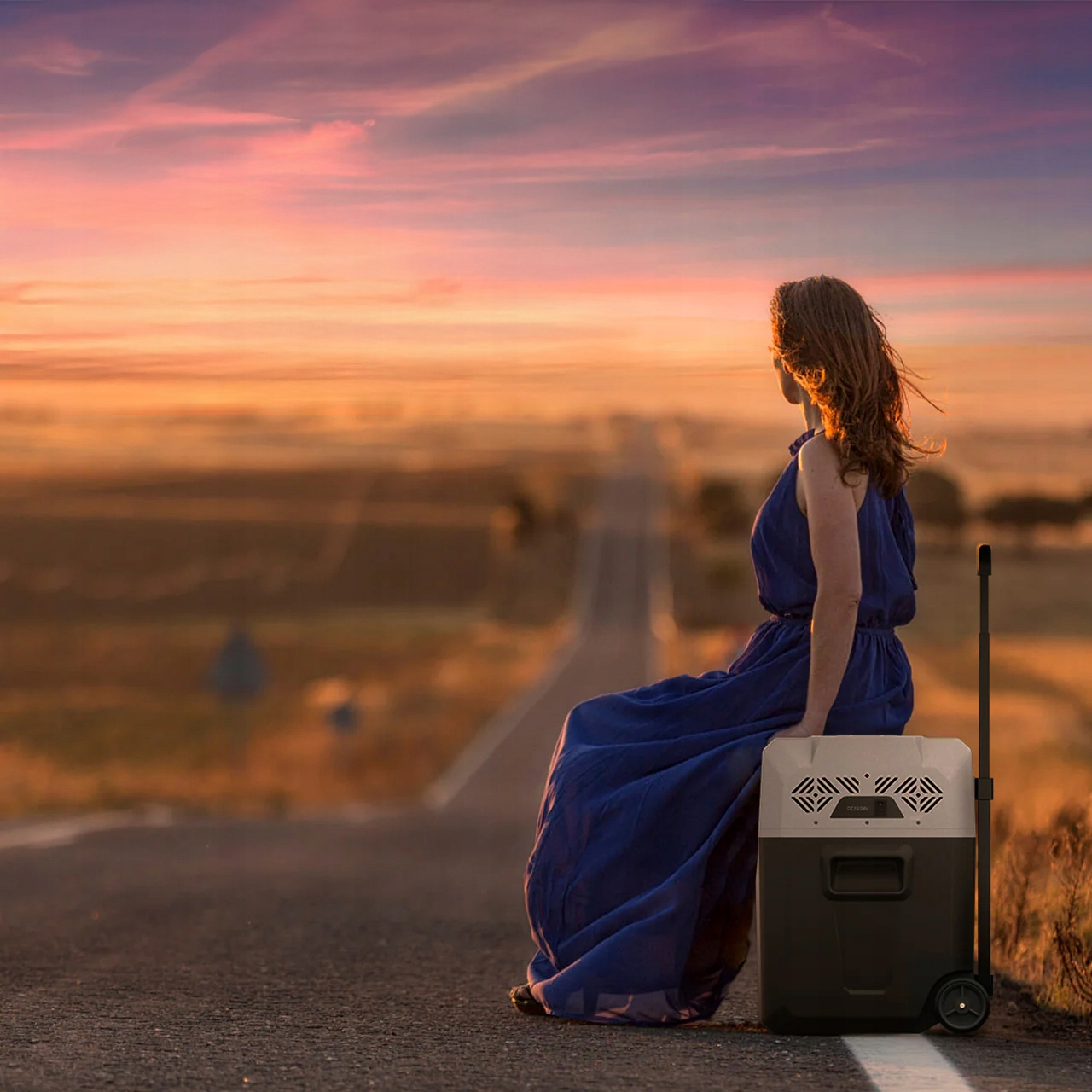 Kobieta w niebieskiej sukience siedząca na drodze, obok chłodziarki, z zachodem słońca w tle.
