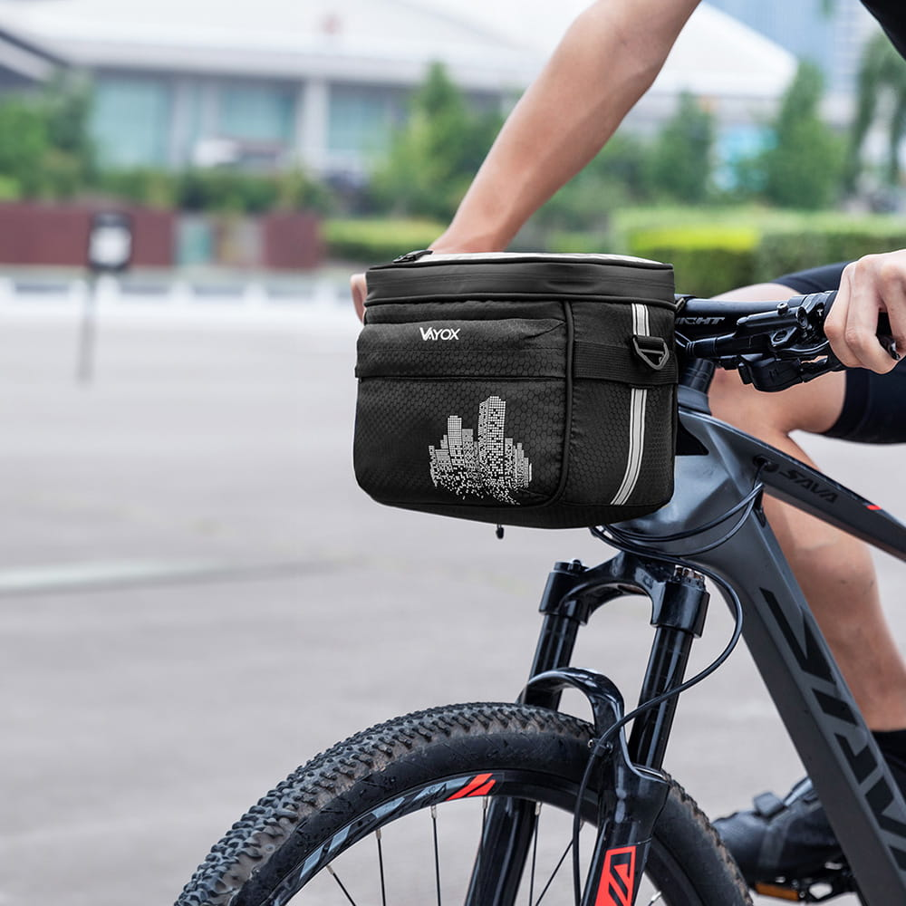 Osoba jadąca na rowerze z czarną torbą rowerową przymocowaną do kierownicy, z sylwetką miasta na torbie.