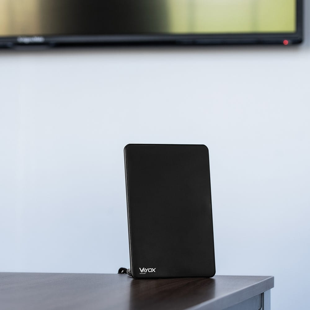 Czarna antena Vayox na stole. W tle telewizor, z białą ścianą.