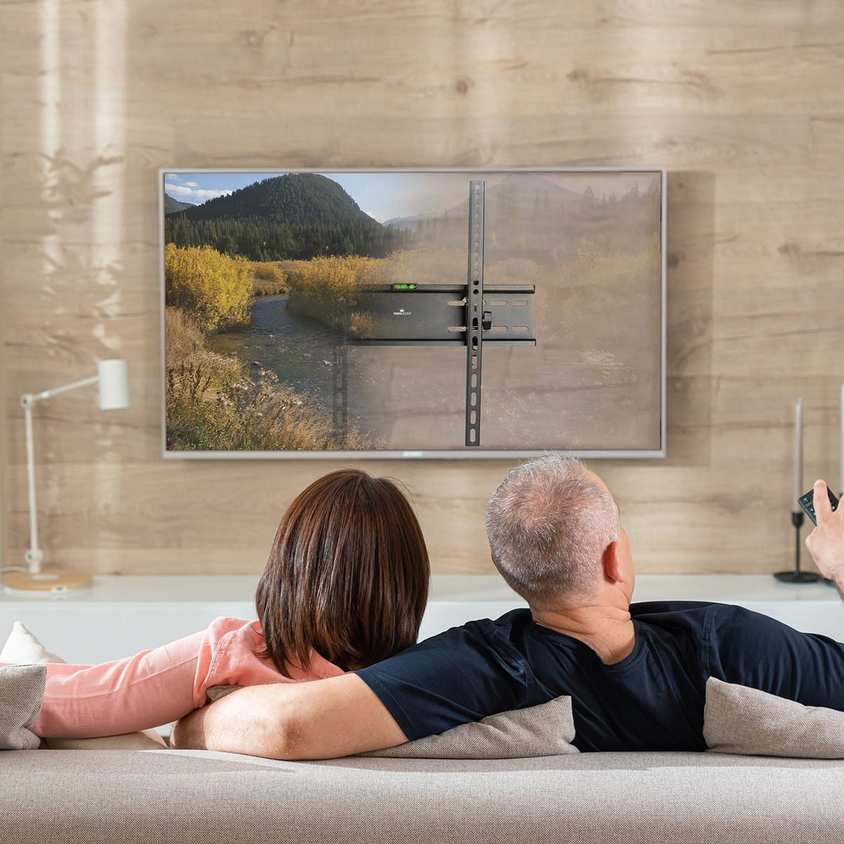 Para siedzi na kanapie oglądając telewizję zamontowaną na ścianie. Uchwyt jest widoczny.