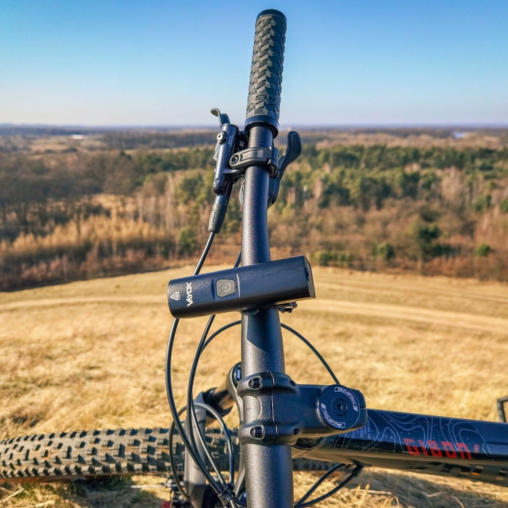 Czarne światło rowerowe na kierownicy. Tło przedstawia pole i drzewa pod błękitnym niebem.