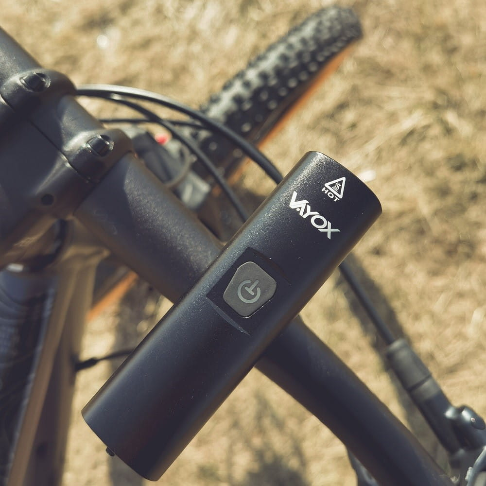 Czarna lampa rowerowa zamontowana na kierownicy roweru, z przyciskiem zasilania, na trawie.