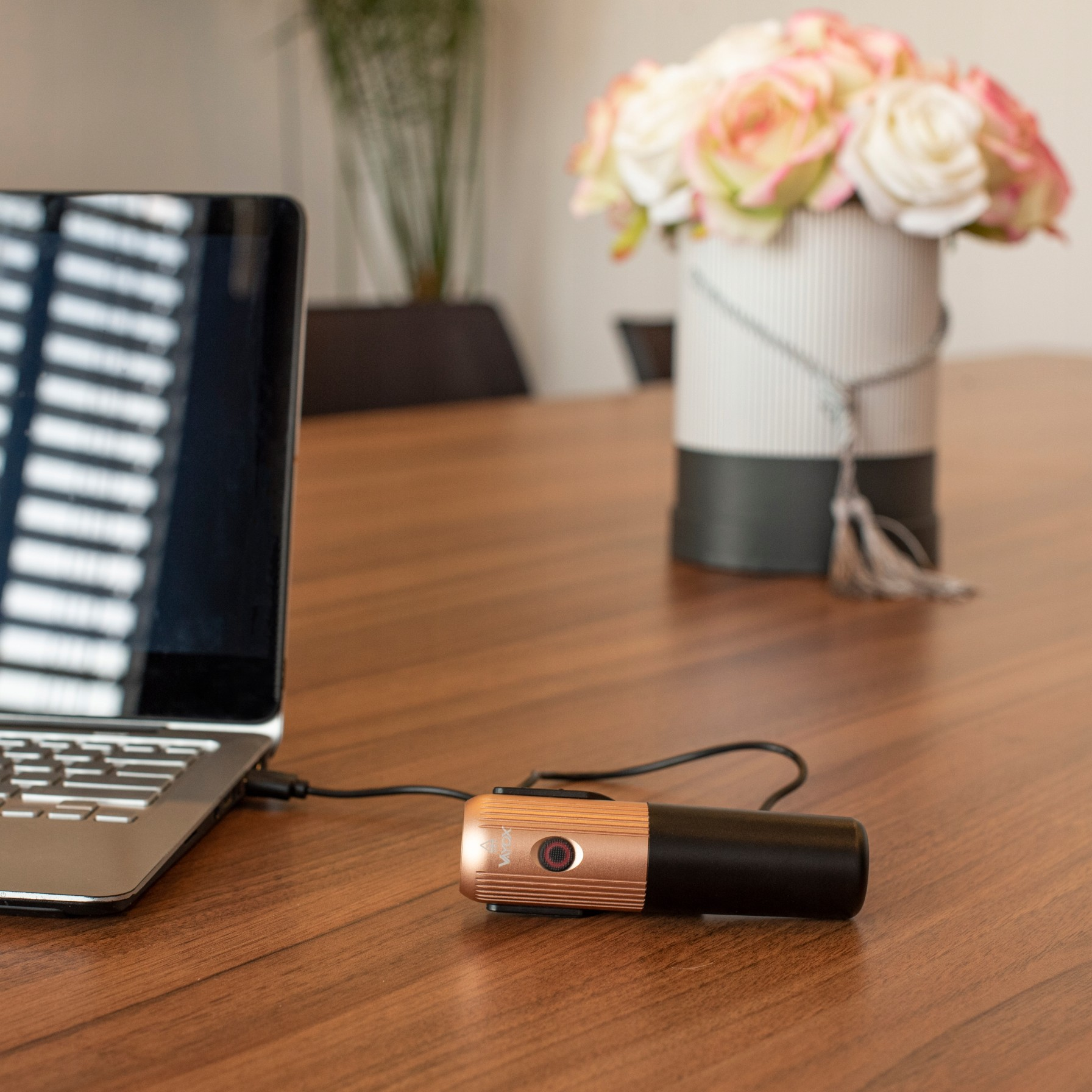 Lampa rowerowa podłączona do laptopa, na drewnianym stole z wazonem kwiatów.