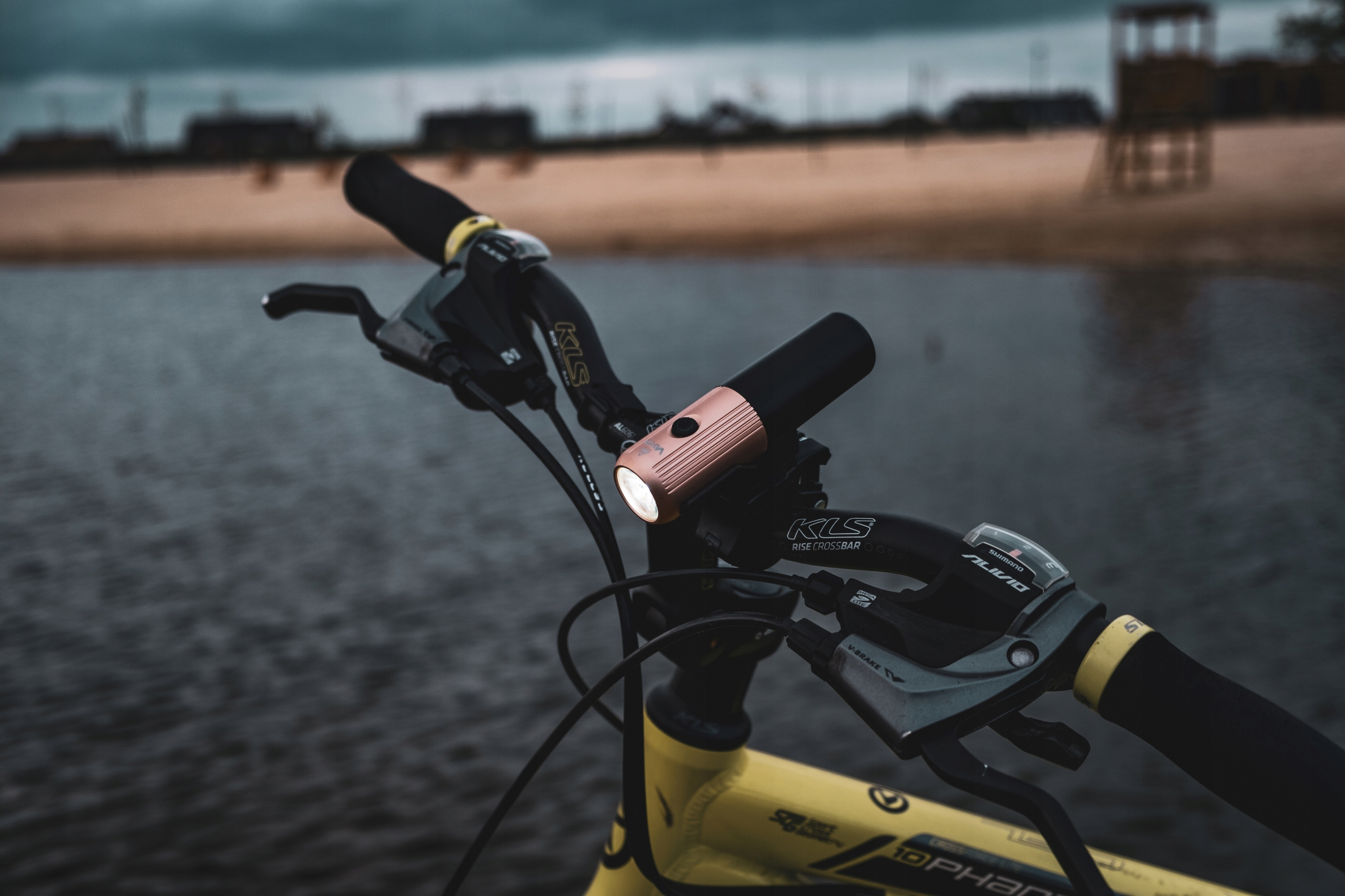 Lampa rowerowa na kierownicy roweru, w pobliżu wody z mostem i zachmurzonym niebem w tle.