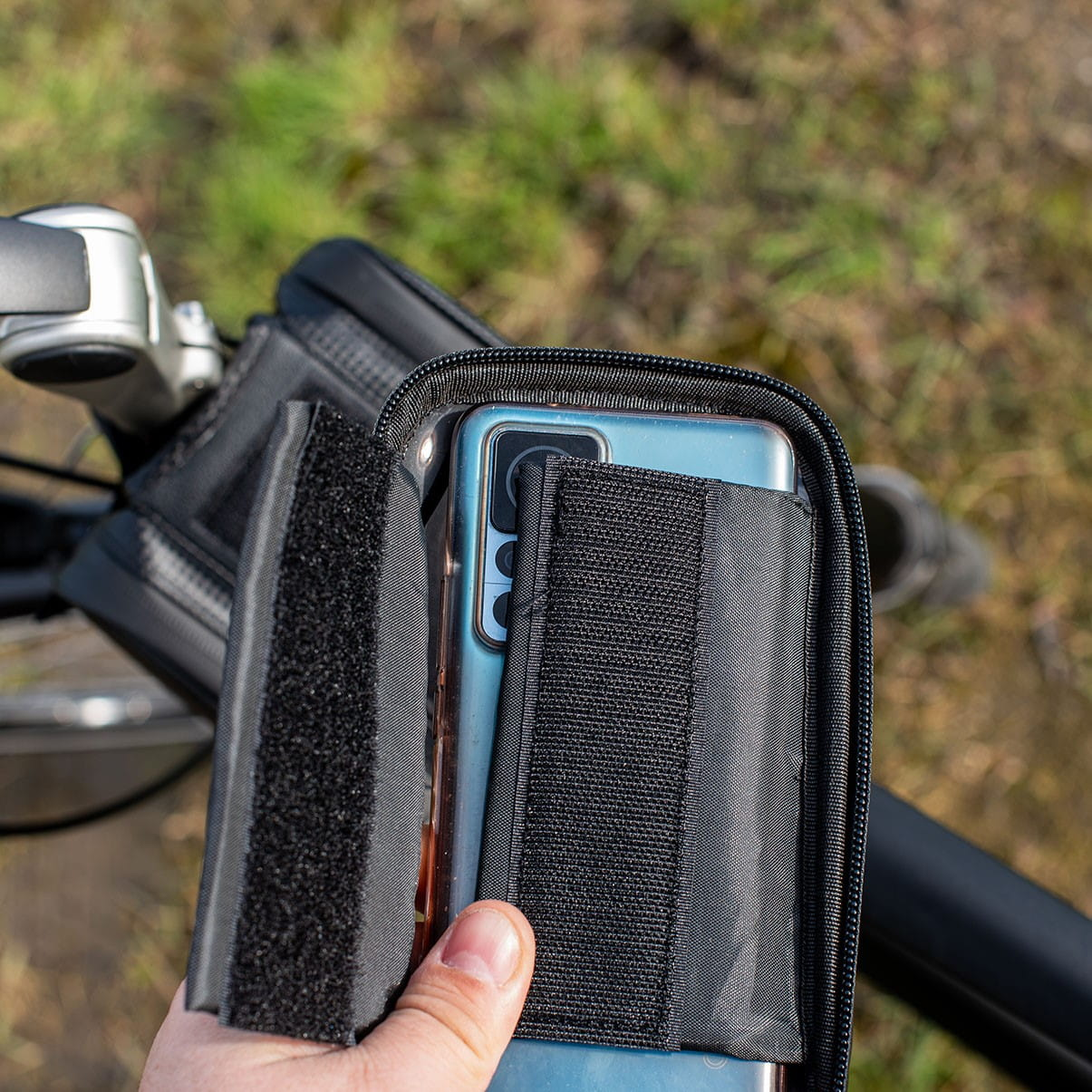 Ręka trzymająca telefon w czarnej torbie rowerowej na kierownicy; zielone tło.