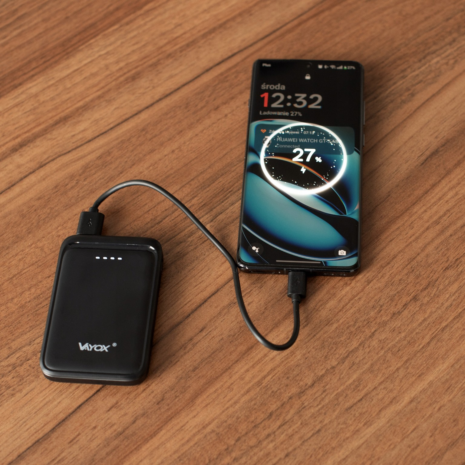 Czarny power bank ładujący smartfona. Telefon pokazuje 27% naładowania. Oba leżą na drewnianym stole.