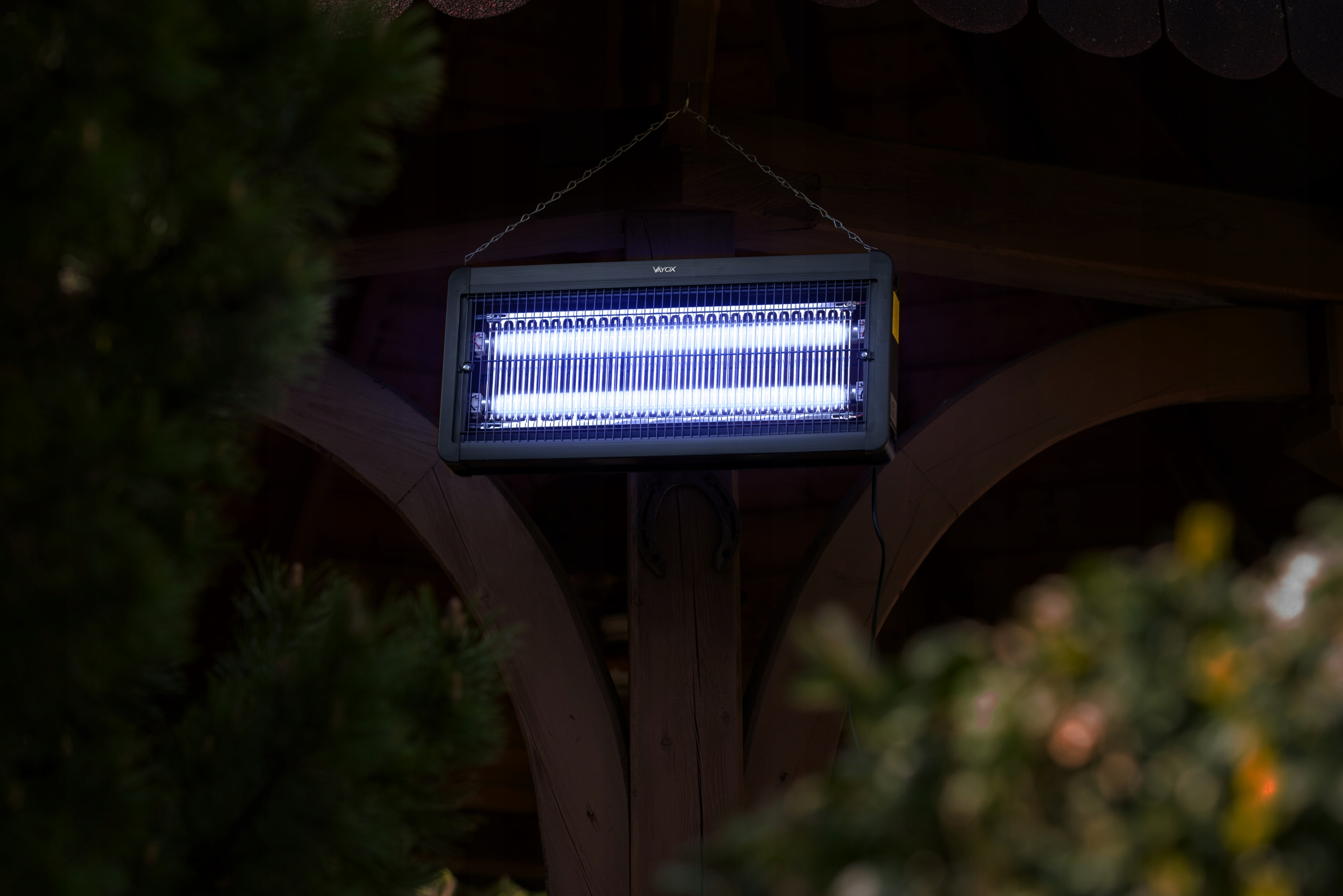 Czarny zabijacz owadów zawieszony na drewnianej konstrukcji. Wewnątrz widoczne niebieskie światło.