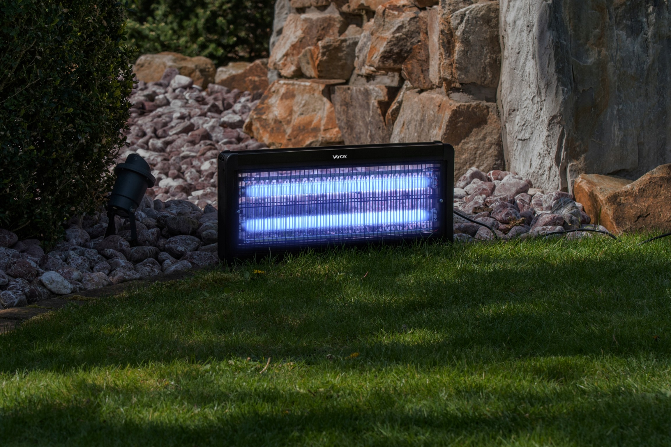 Czarny zabijacz owadów na zielonej trawie, w tle kamienna ściana. Emituje niebieskie światło.