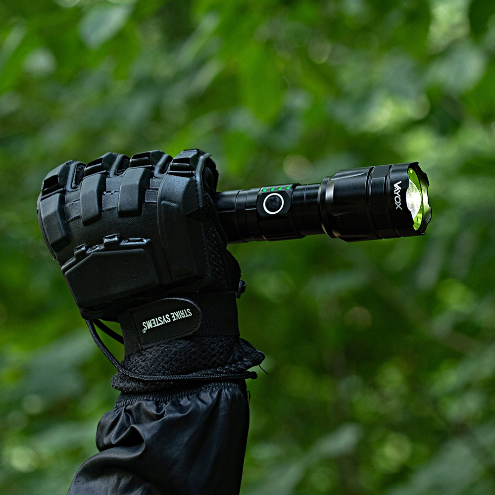 Ręka w rękawicy trzymająca czarną latarkę, z zielonym światłem, na zewnątrz w lesie.