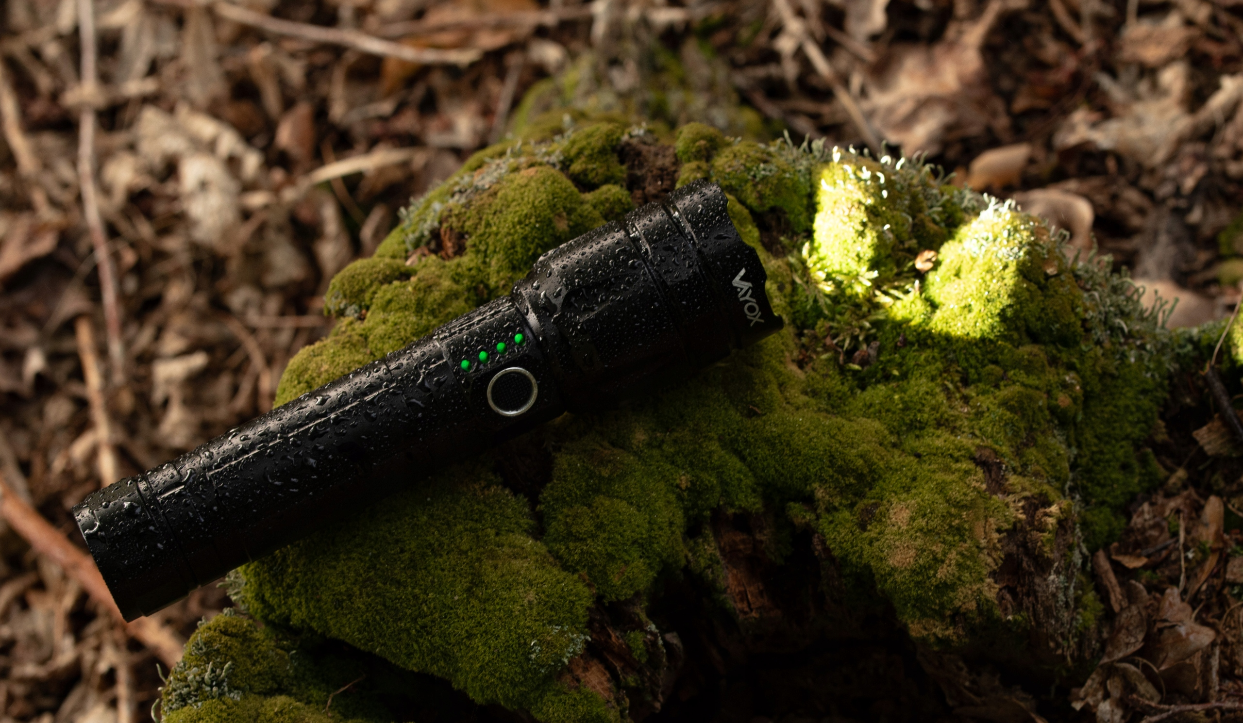 Czarna latarka leżąca na omszałej ziemi. Latarka jest mokra. Świecą zielone wskaźniki.