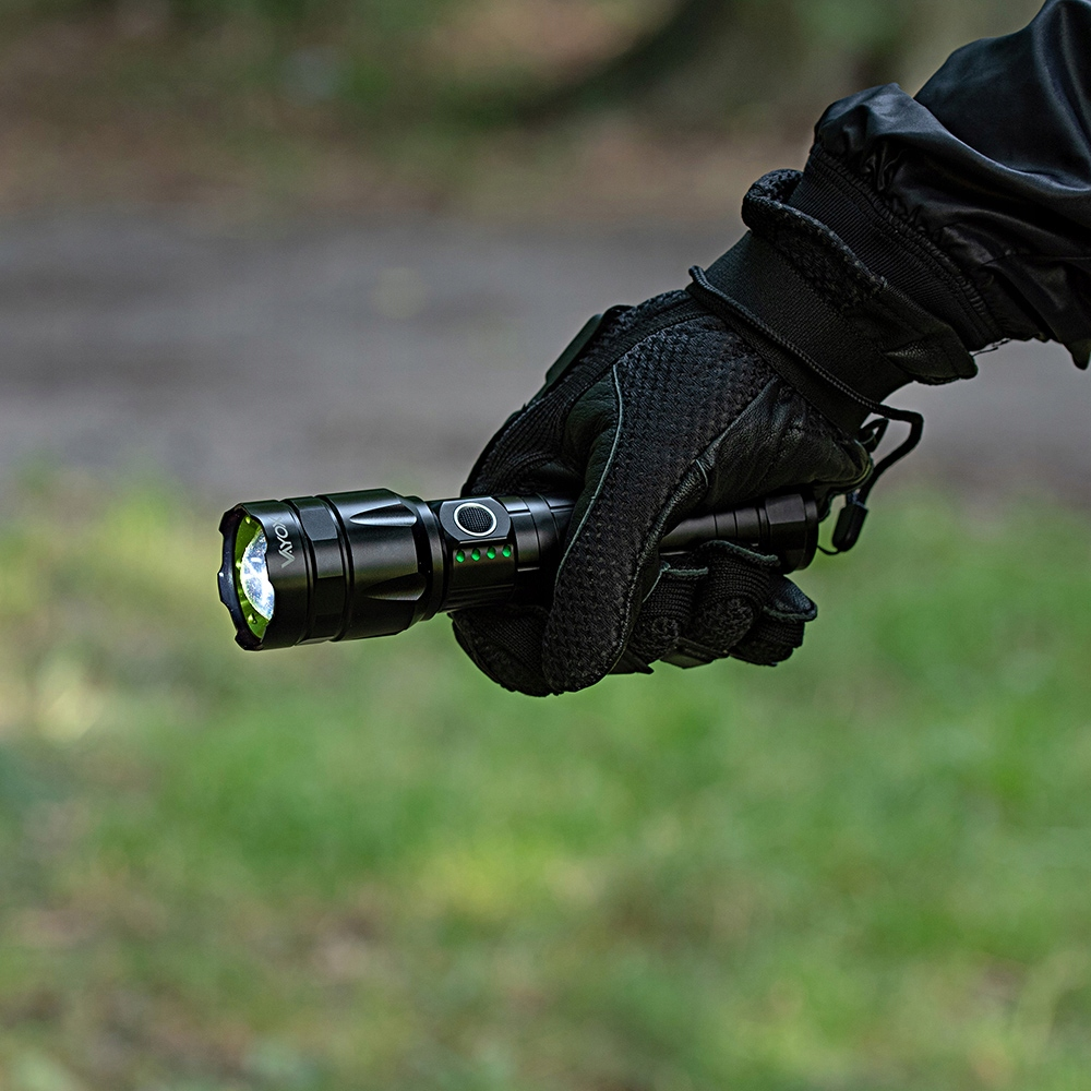 Ręka w rękawicy trzymająca czarną latarkę. Latarka jest włączona, emitując jasne, białe światło.