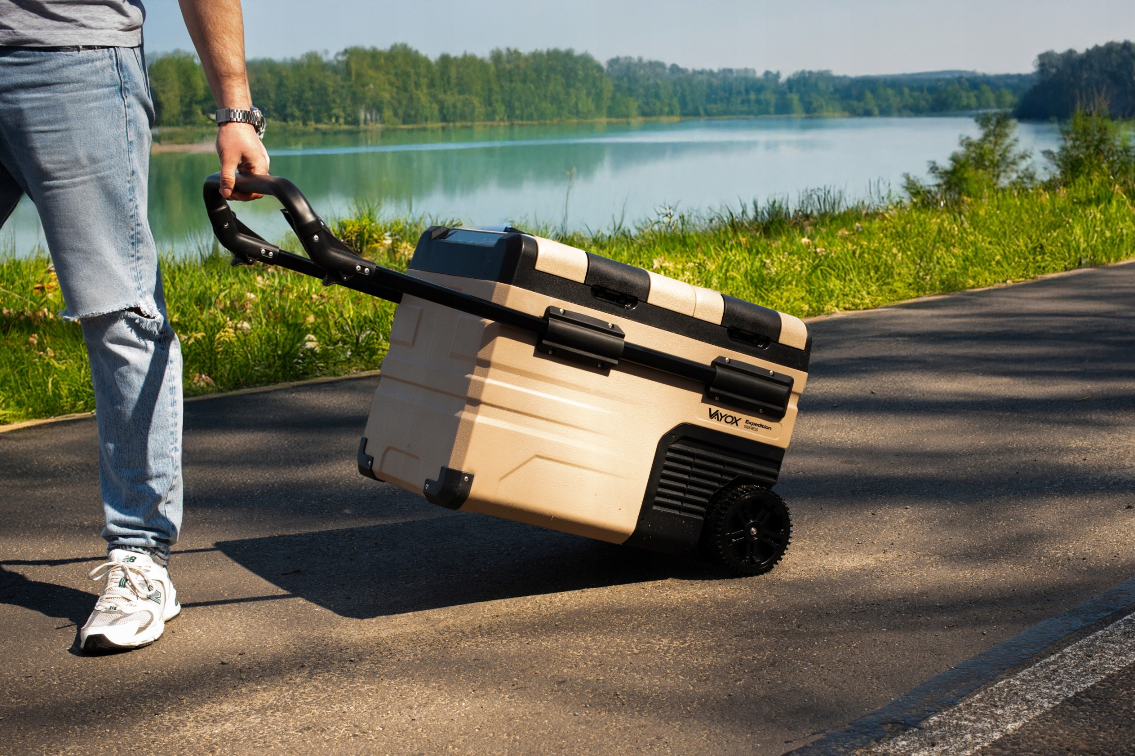 Osoba ciągnąca beżową chłodziarkę na kółkach po utwardzonej ścieżce w pobliżu jeziora.