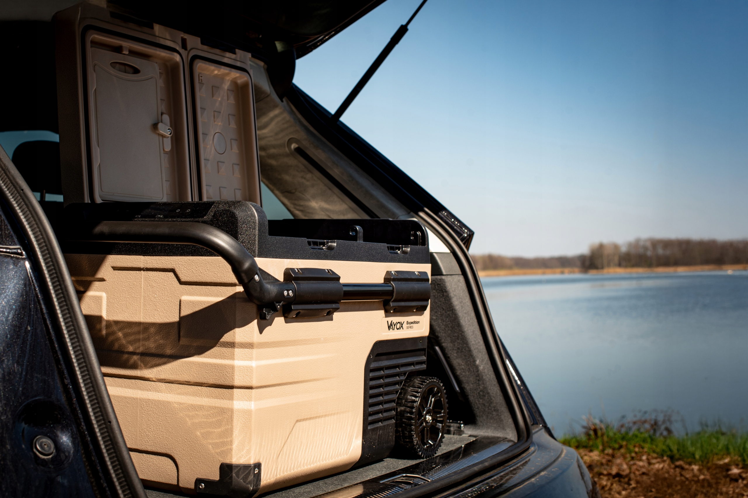 Beżowa chłodziarka w bagażniku samochodu, z otwartą pokrywą ukazującą zawartość, w pobliżu jeziora.