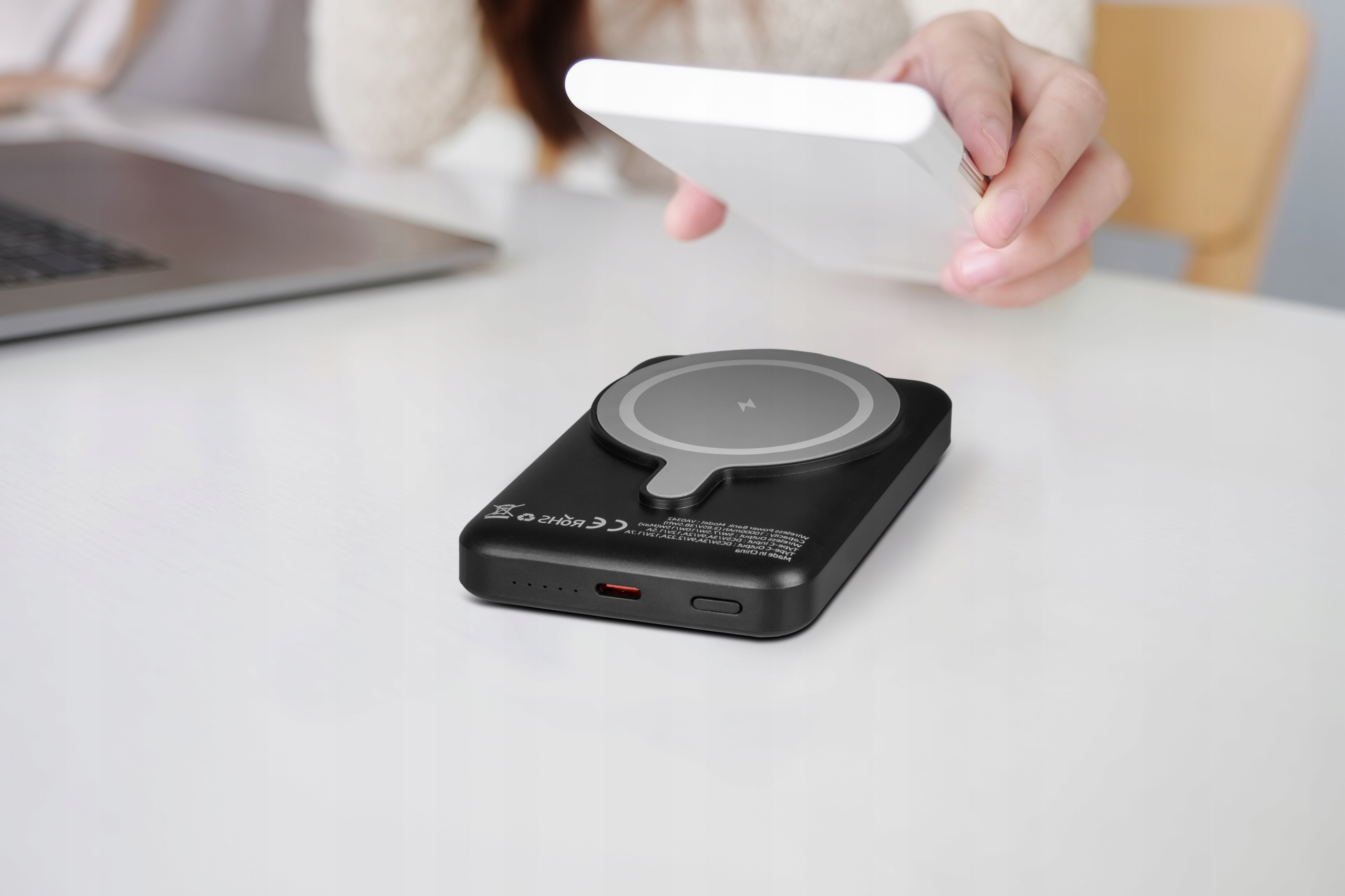 Ręce trzymają białą podkładkę ładującą. Czarny power bank leży na białym stole.