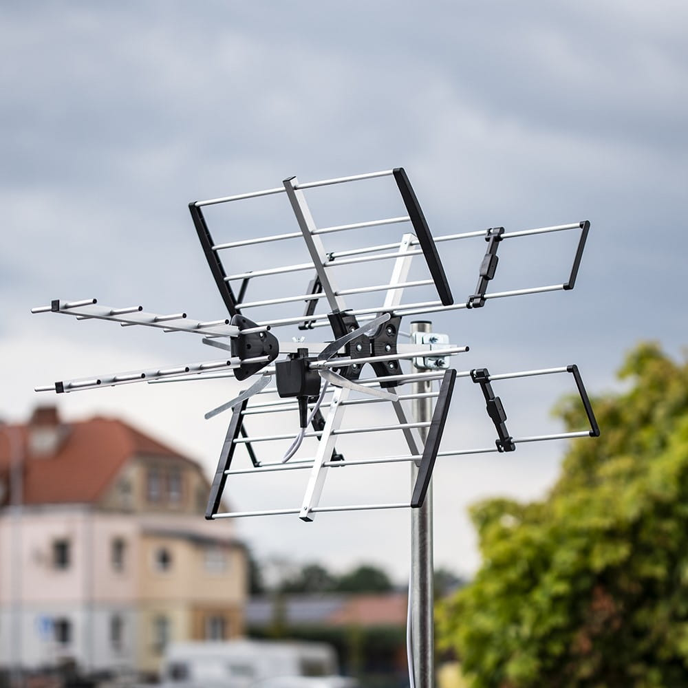 Antena telewizyjna zewnętrzna zamontowana na słupie. Tło pokazuje zachmurzone niebo, budynki i drzewa. Srebrny i czarny.