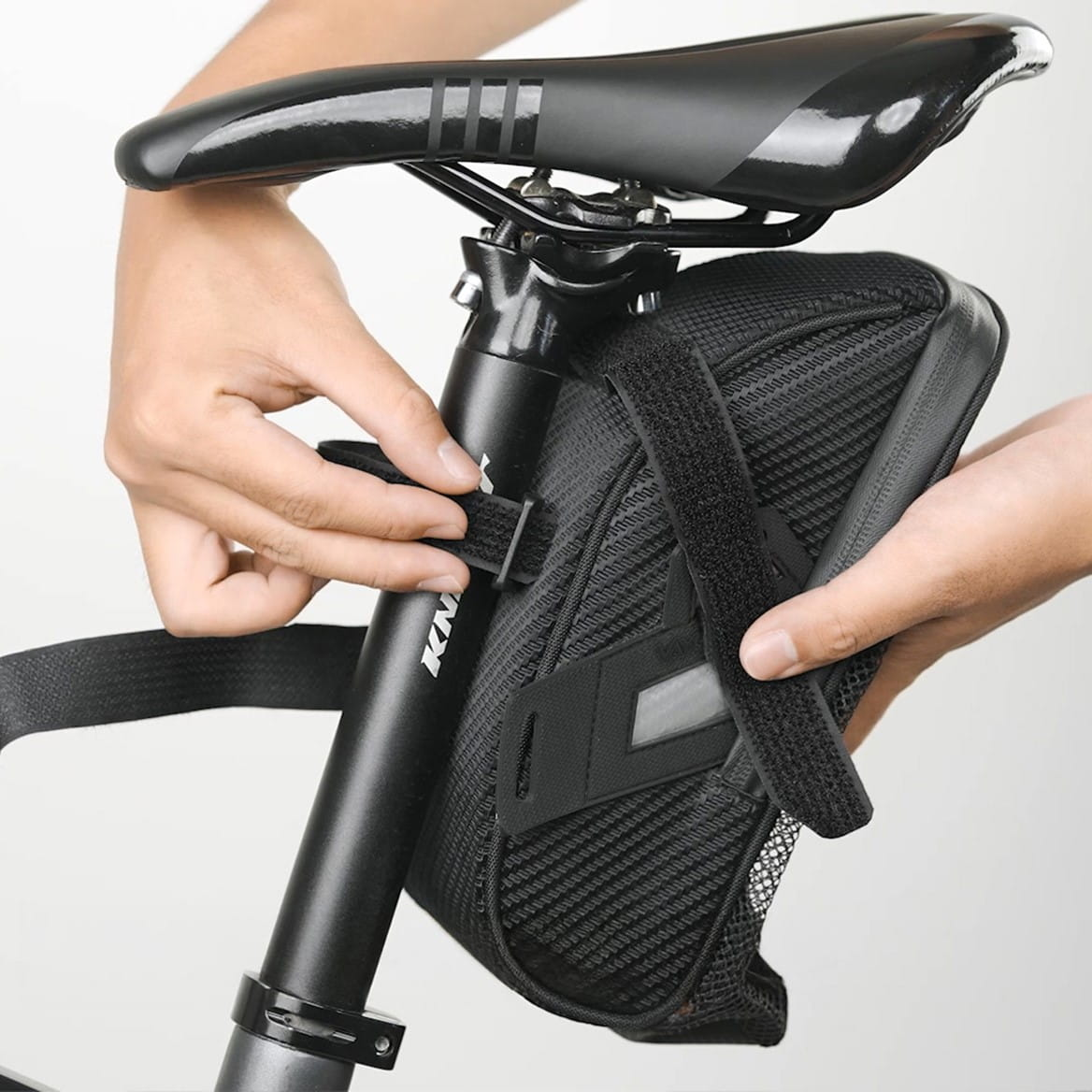 Osoba mocująca czarną torbę rowerową do sztycy podsiodłowej. Torba jest czarna z odblaskowym paskiem.