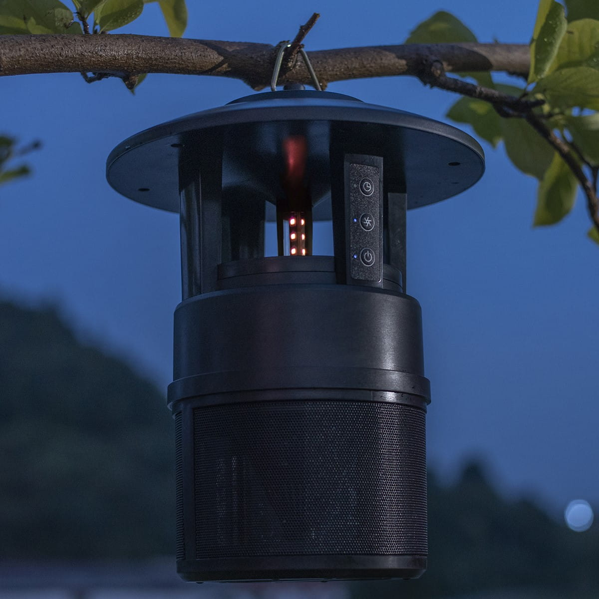 Czarny głośnik/światło zewnętrzne, wiszące na gałęzi w nocy, z błękitnym niebem.