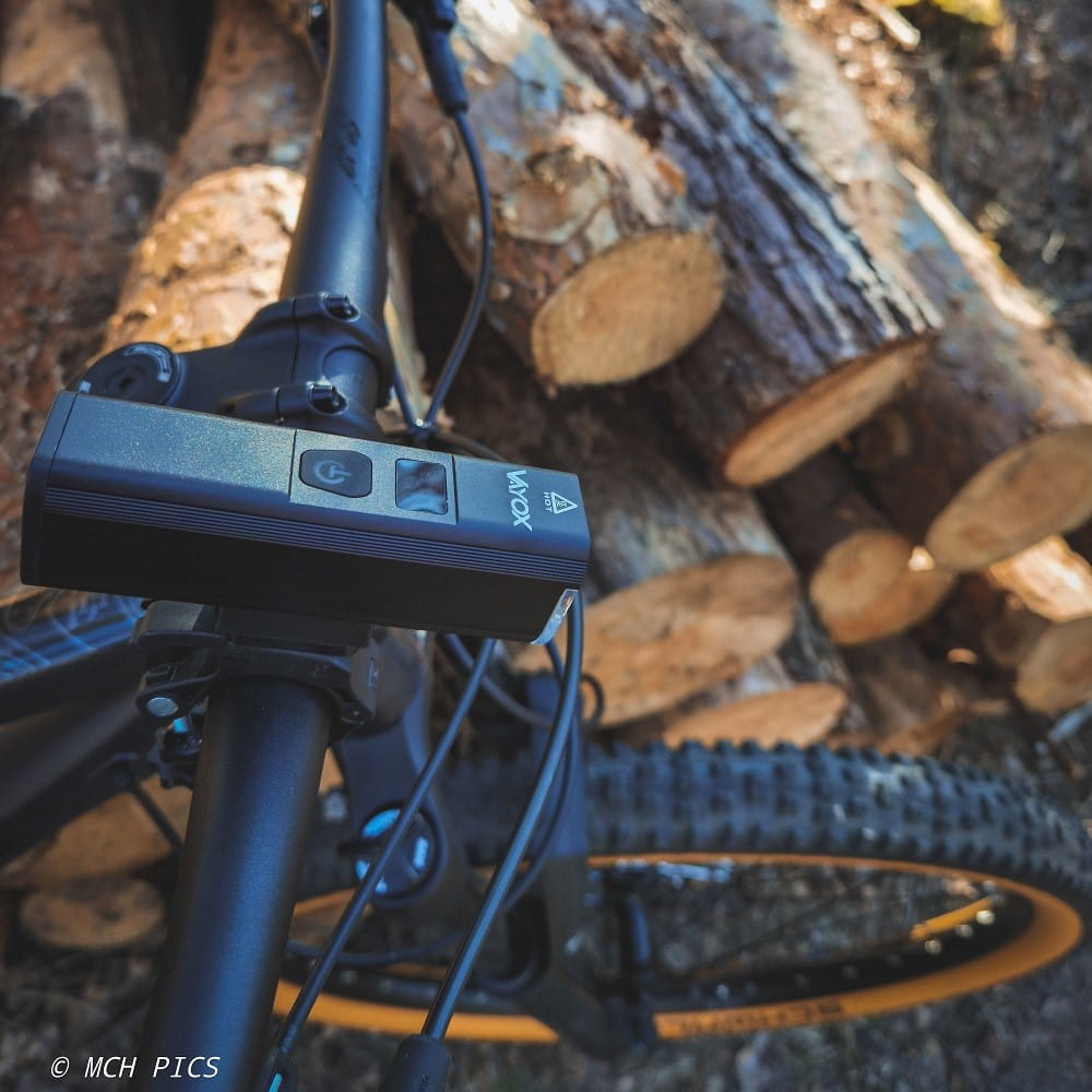 Czarna lampka rowerowa zamontowana na rowerze. Tło przedstawia kłody drewna.