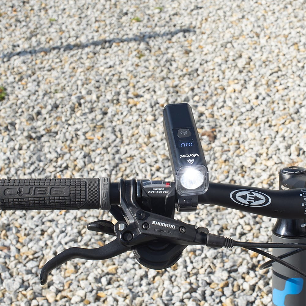 Czarna lampka rowerowa na kierownicy. Tło to żwir. Komponenty Shimano Deore.