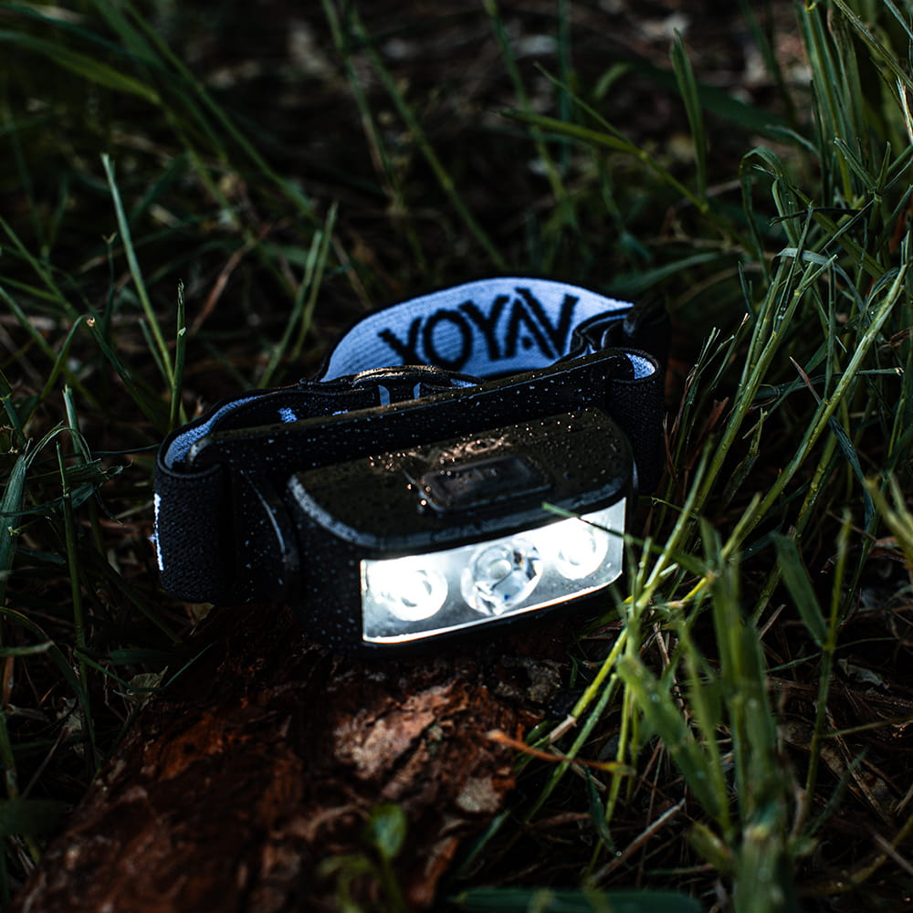 Czarna lampa czołowa z trzema jasnymi światłami na ciemnym tle trawy i kłody.