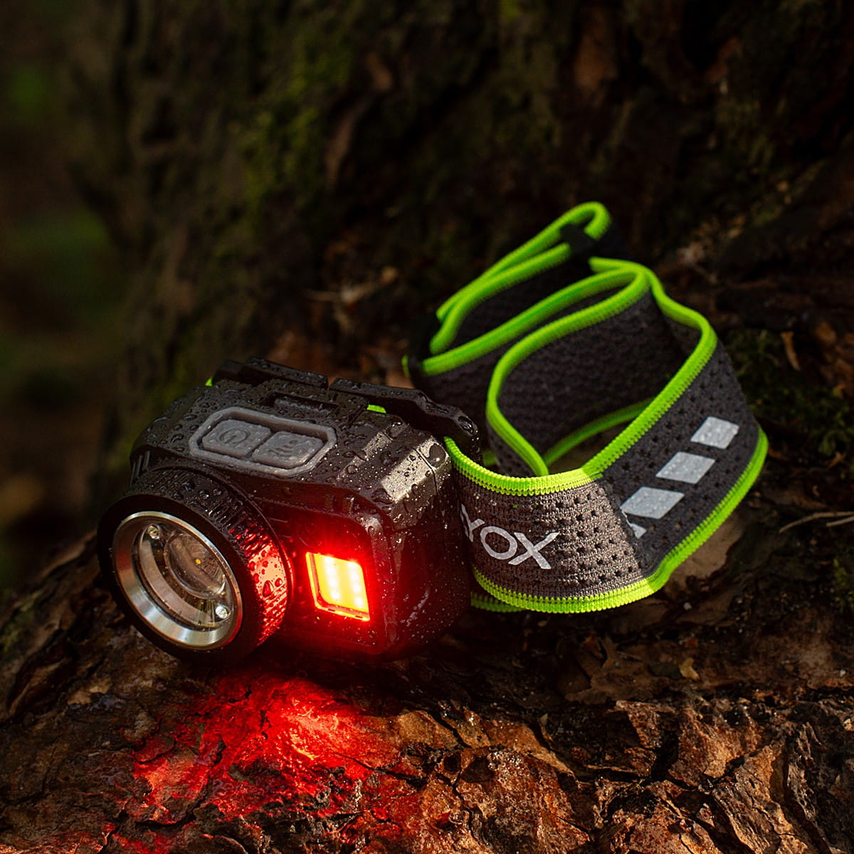 Czarna latarka czołowa z czerwonym światłem, na pniu drzewa, z zielono-szarym paskiem.