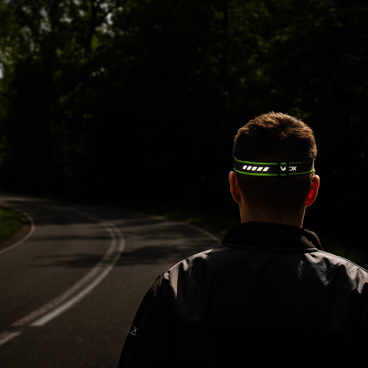 Osoba z zielono-białą opaską na głowie idzie drogą, ciemny las w tle.