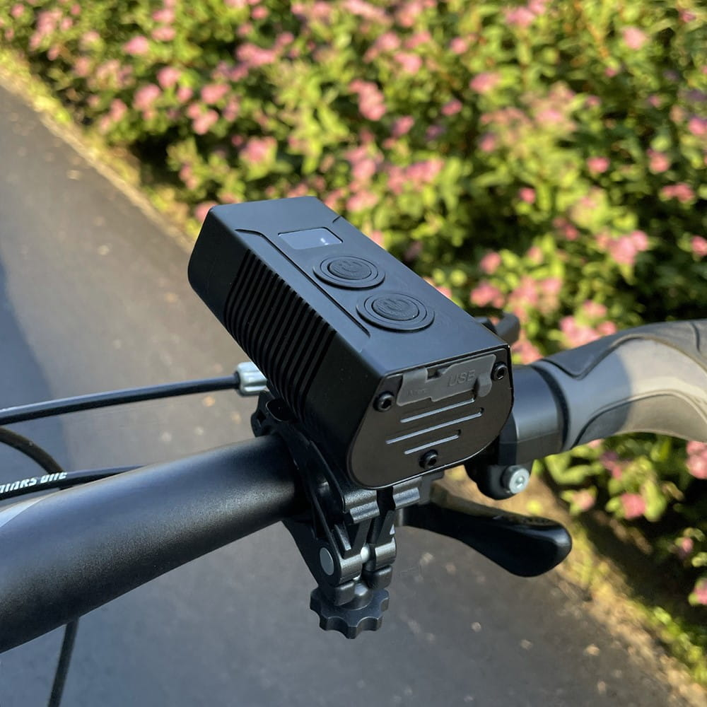 Czarne światło rowerowe zamontowane na kierownicy. Widoczne są dwa przyciski. Tło jest rozmyte.