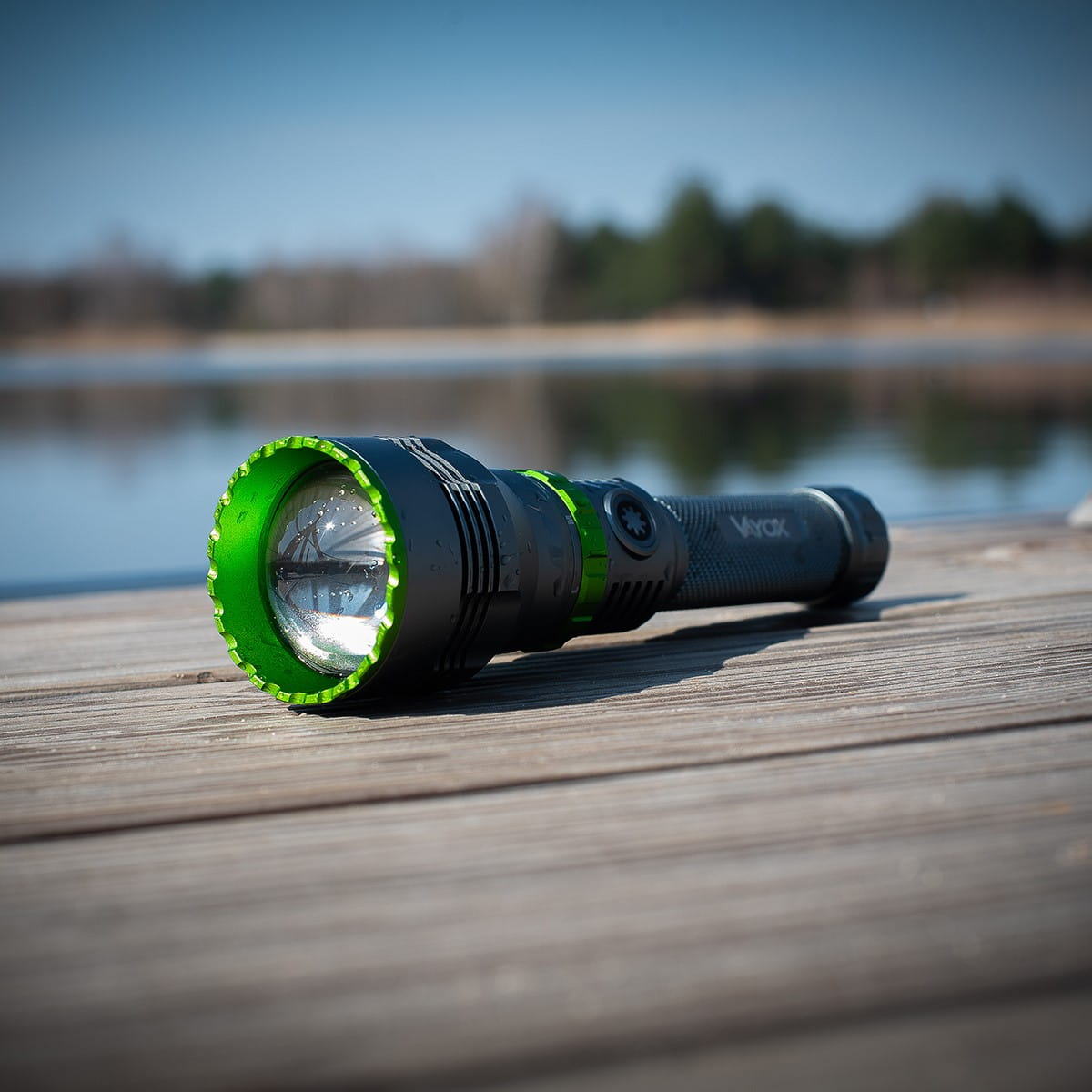 Czarna i zielona latarka na drewnianym pomoście. Światło świeci jasno. Widoczne jest jezioro.