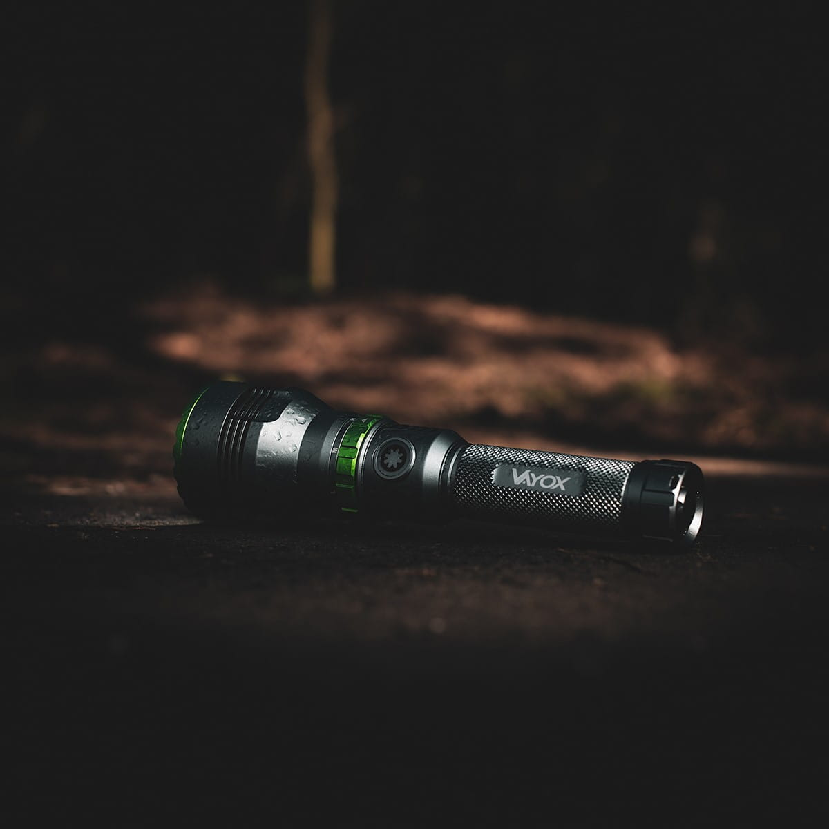 Czarna i zielona latarka leżąca na ciemnej leśnej podłodze z lekko rozmytym tłem.