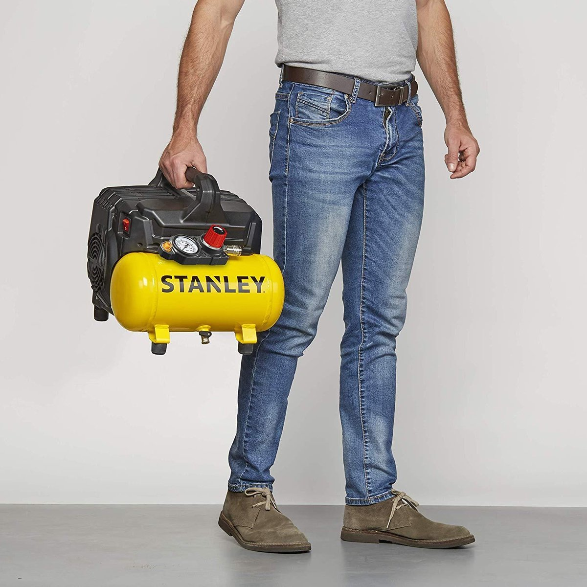 Een man in een spijkerbroek en een grijs overhemd die een geel-zwarte luchtcompressor met een handvat draagt.