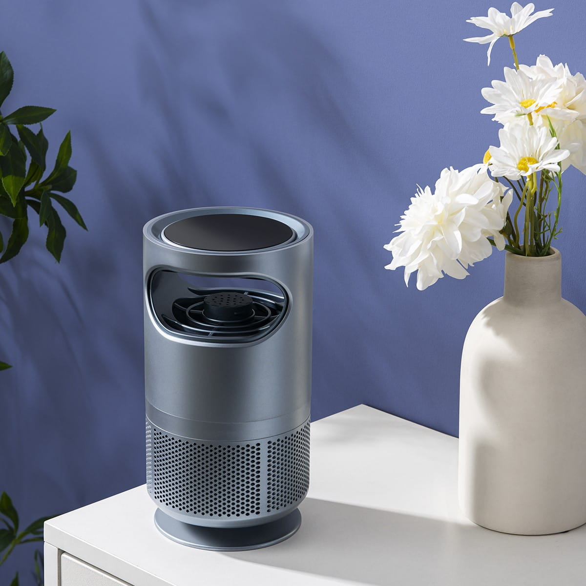 Srebrna lampa owadobójcza na białym stole z kwiatami w wazonie na tle niebieskiej ściany.