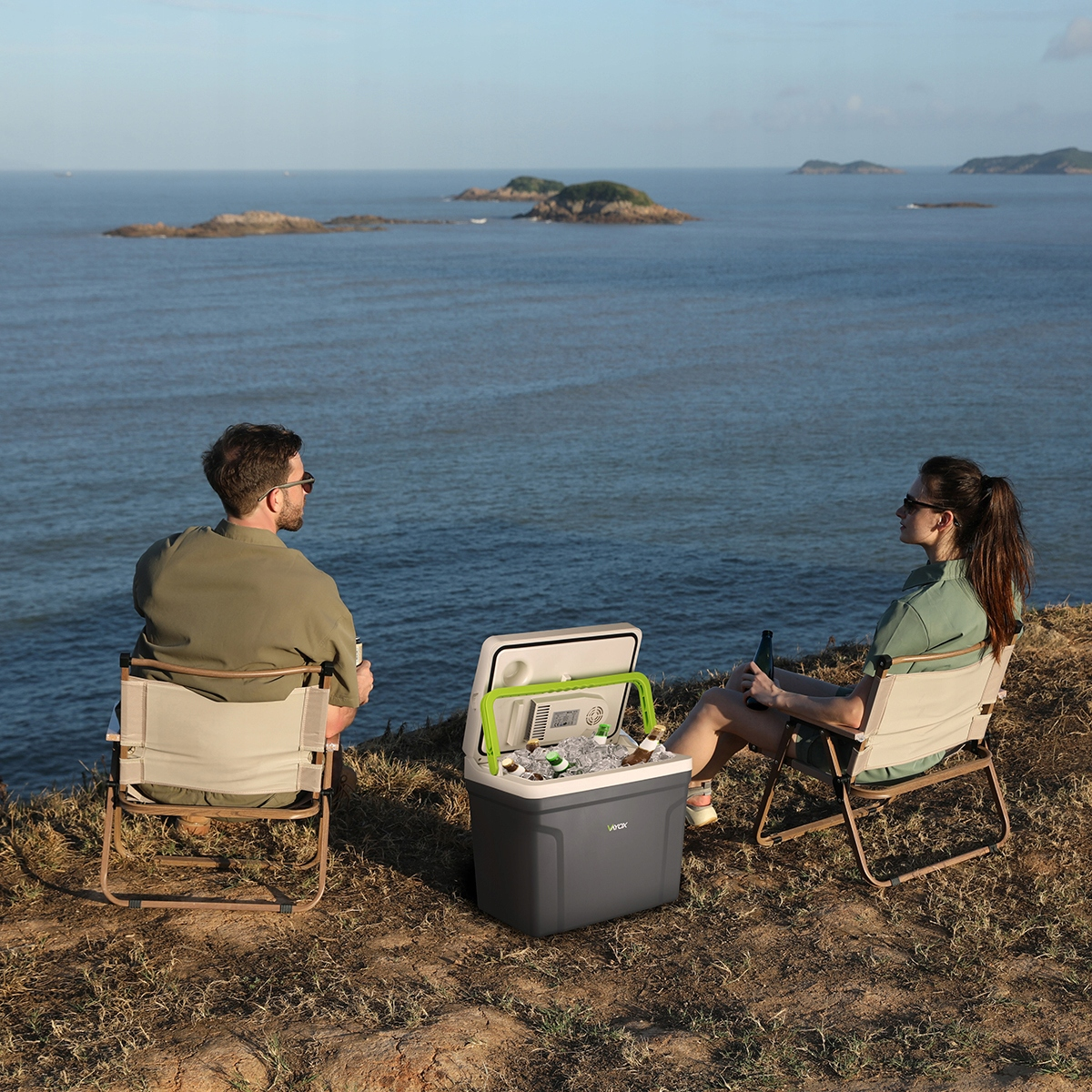 Dwie osoby siedzące na krzesłach z chłodziarką między nimi. Tłem jest widok na morze.